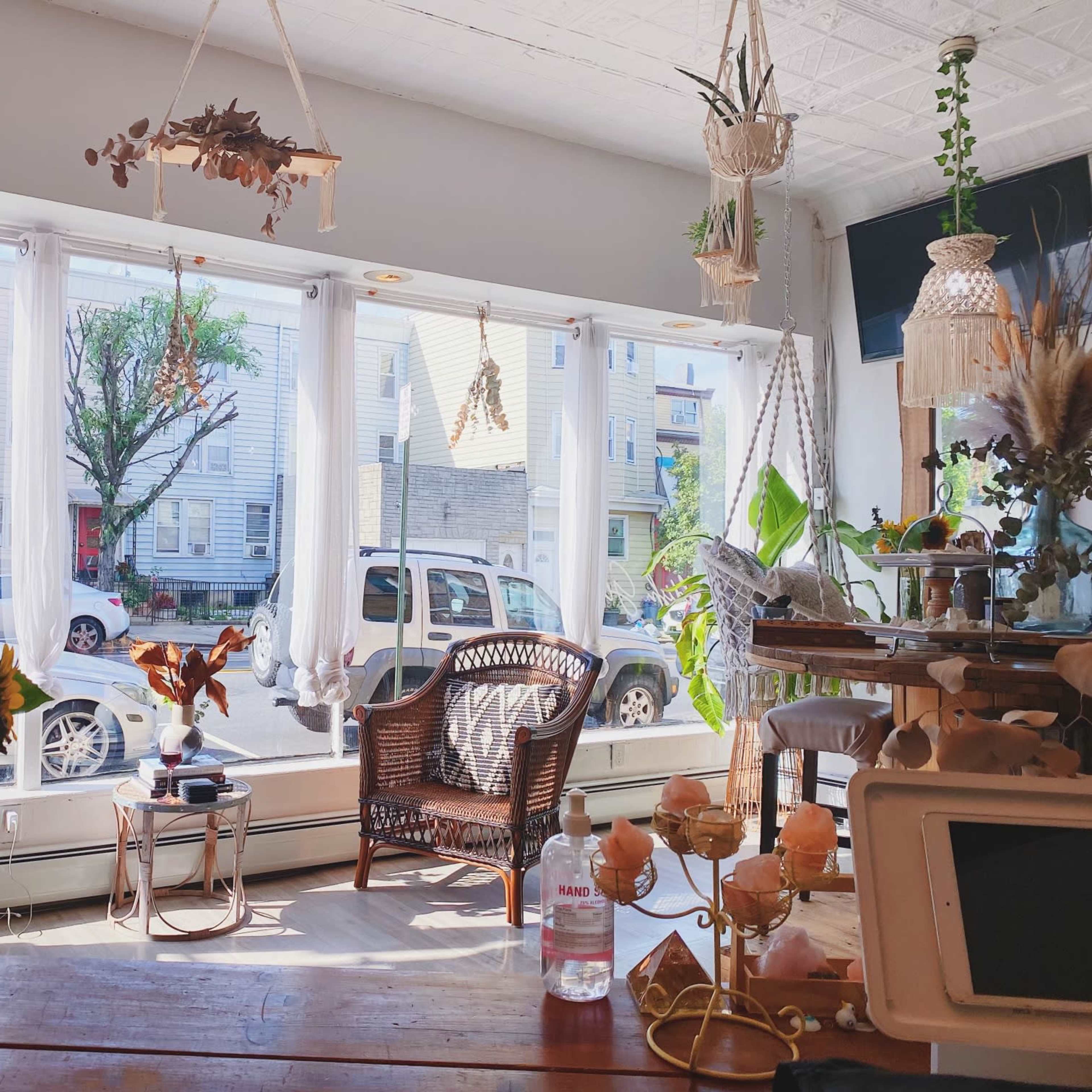 A bright interior of a cafe with wicker chairs and a wooden table, featuring plants and sunlight streaming through large windows overlooking a street.