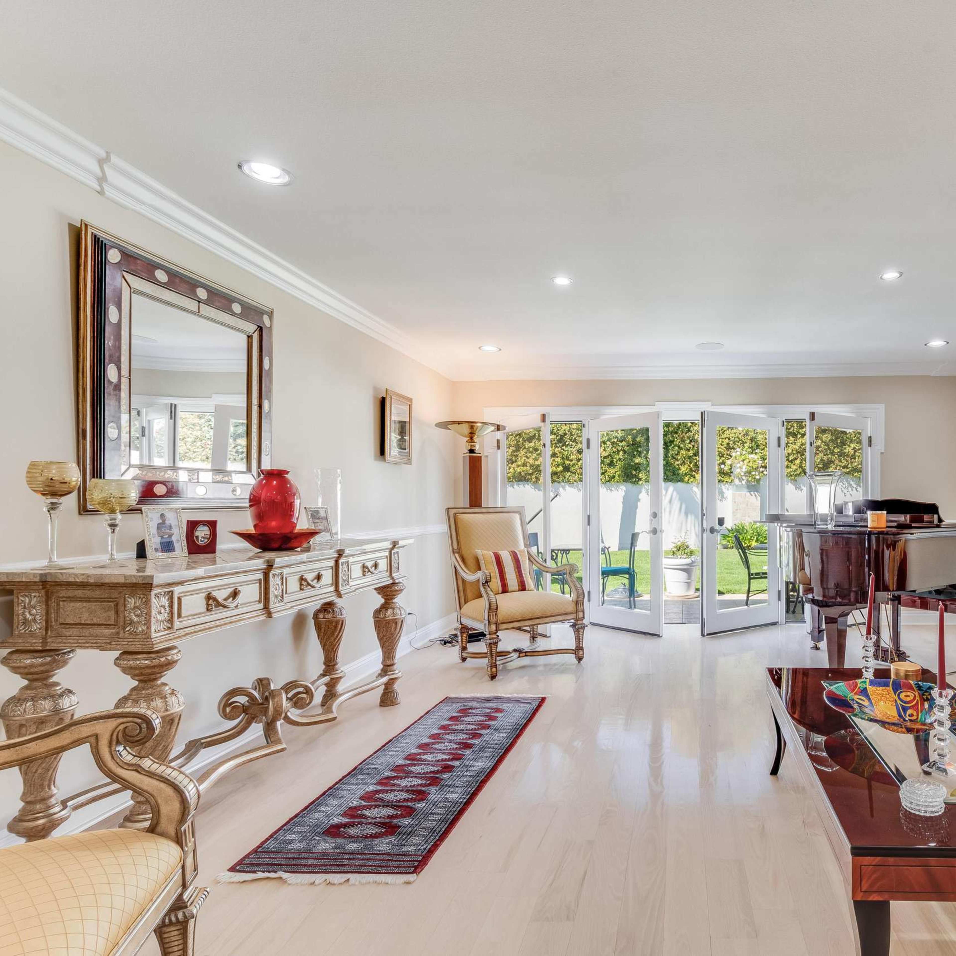 A spacious living room with a red sofa, a wooden piano, a decorative console table, and large glass doors opening to a garden.
