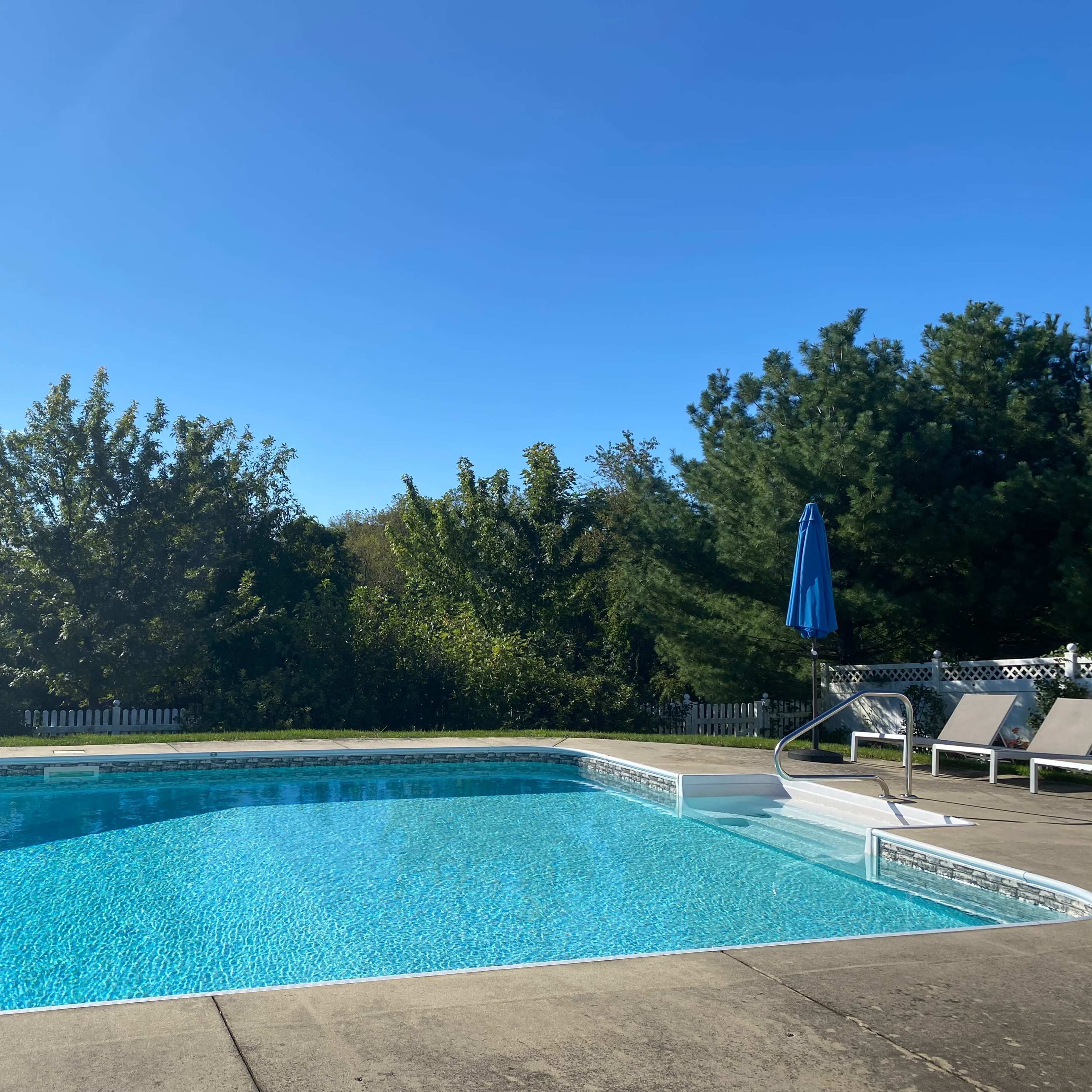 A clear swimming pool surrounded by green trees and lawn chairs under a blue umbrella.
