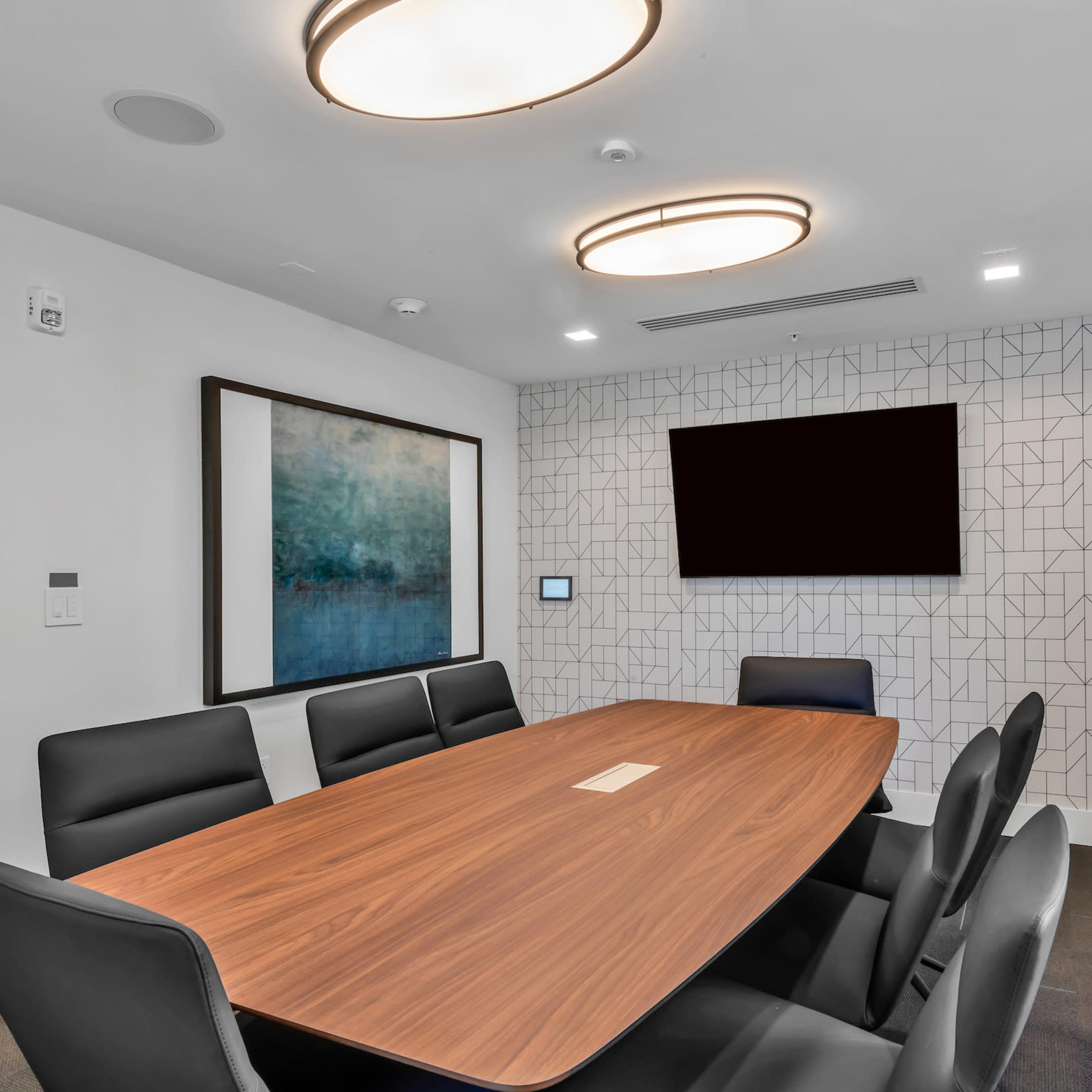 A modern conference room features a large wooden table surrounded by black chairs, with a wall-mounted screen and geometric patterned decor.