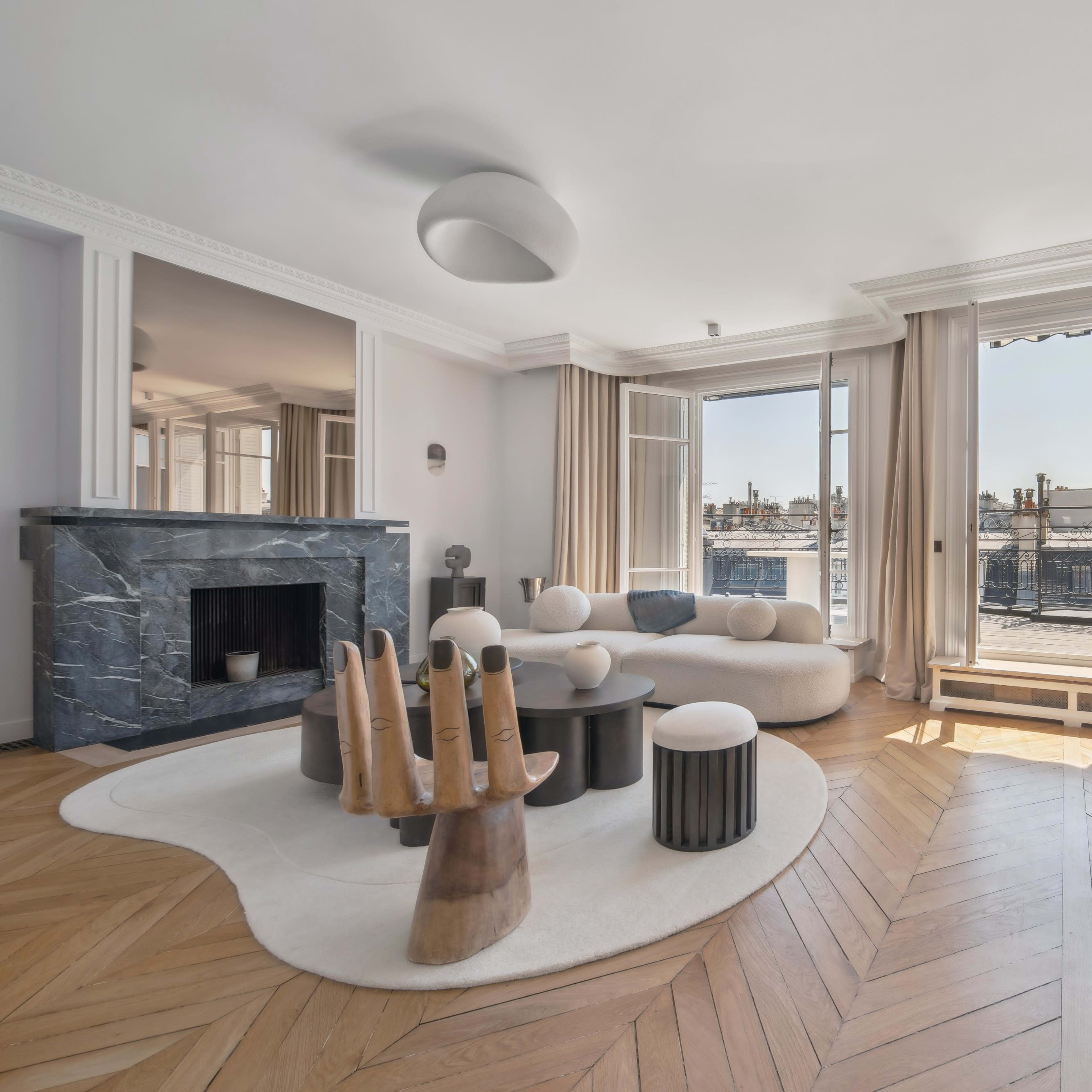 The spacious living room features a marble fireplace, modern furniture, and large windows overlooking a cityscape.