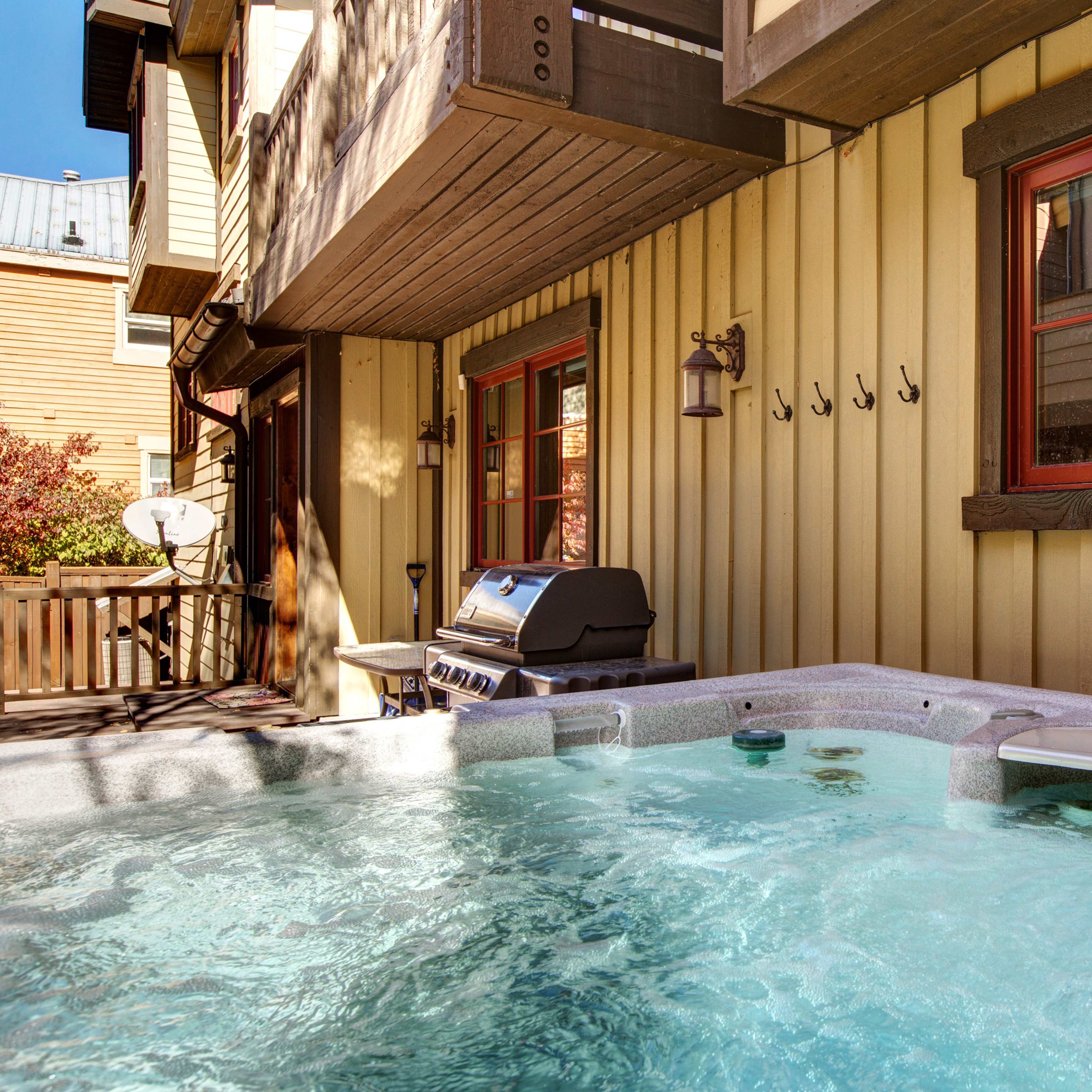 A hot tub situated next to a wooden building with two red-trimmed windows and a barbecue grill nearby.