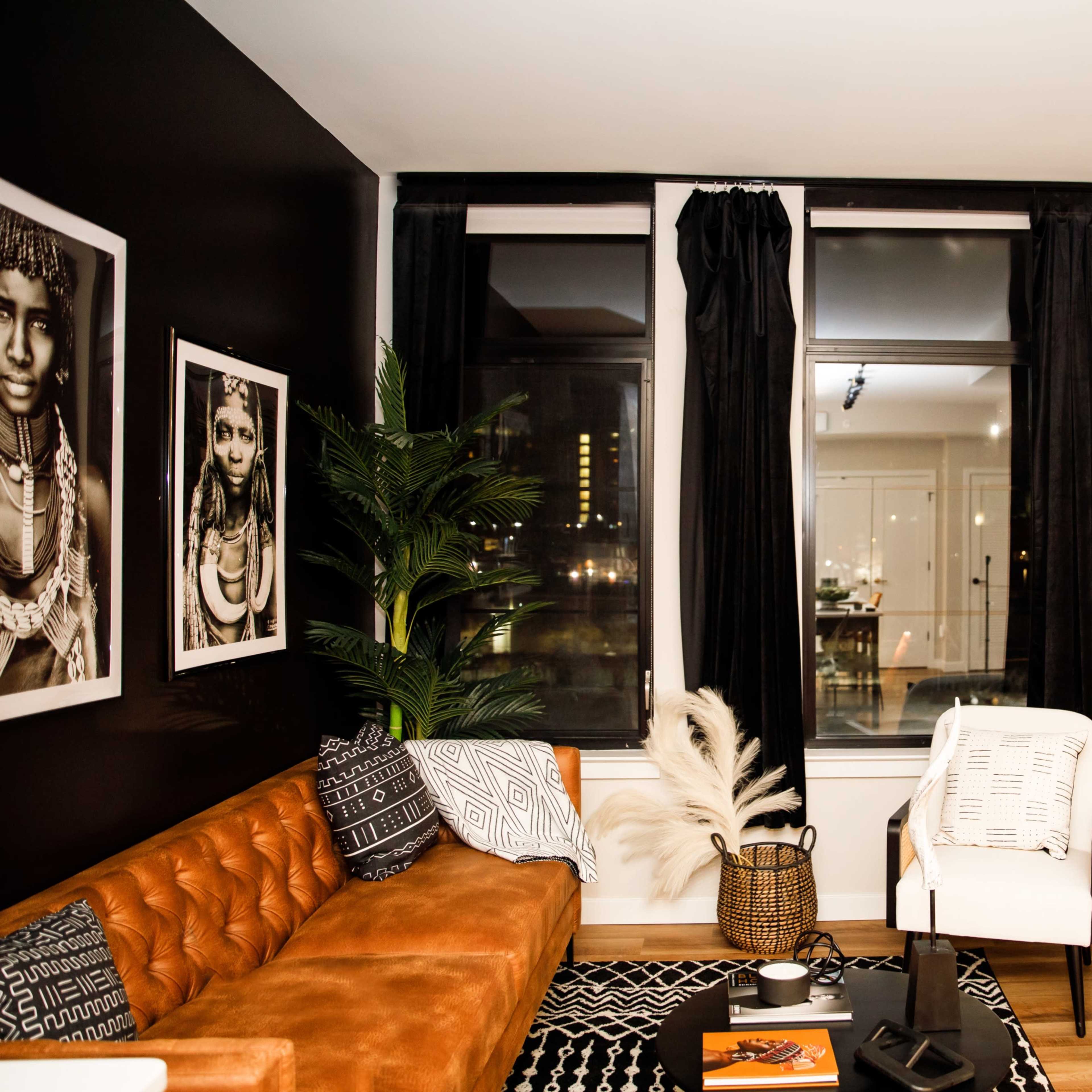 A modern living room with a brown leather sectional sofa, a white chair, large windows with black curtains, and framed artwork on the walls.
