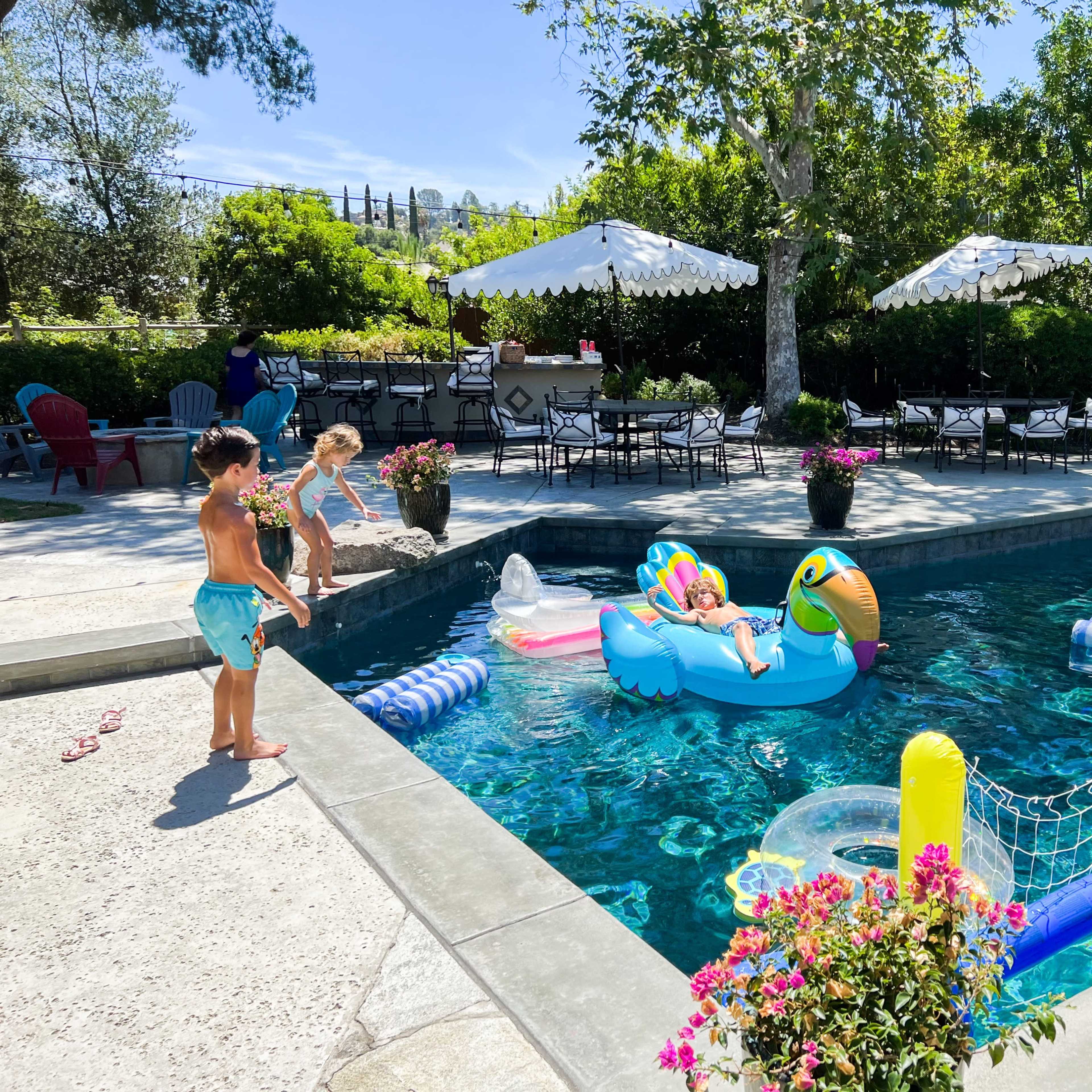 A child stands on the edge of a pool while another child plays with inflatable toys in the water, surrounded by chairs and tables in a sunny backyard.