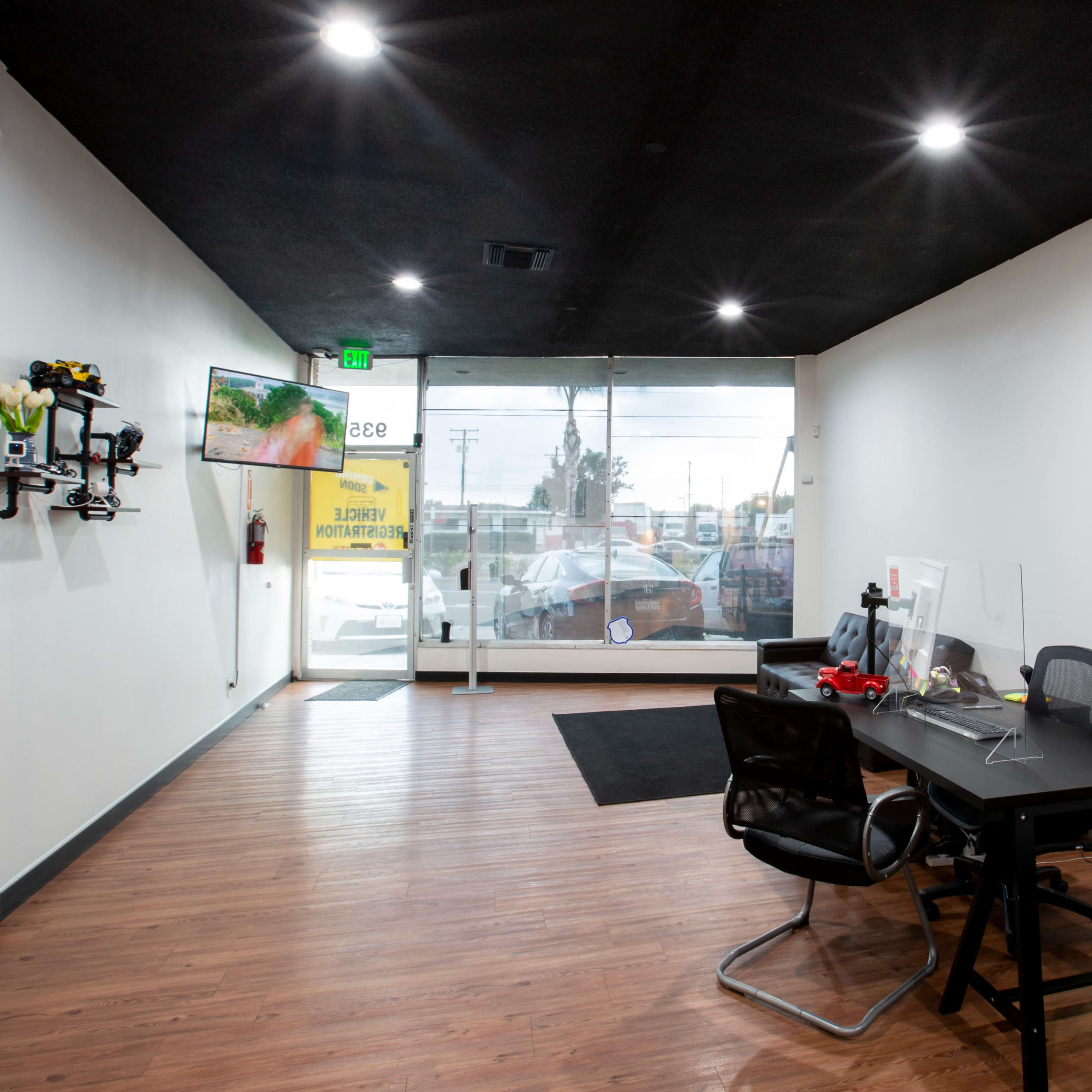 An empty commercial space features a black ceiling, wood flooring, a desk with chairs, and a large window at the entrance.