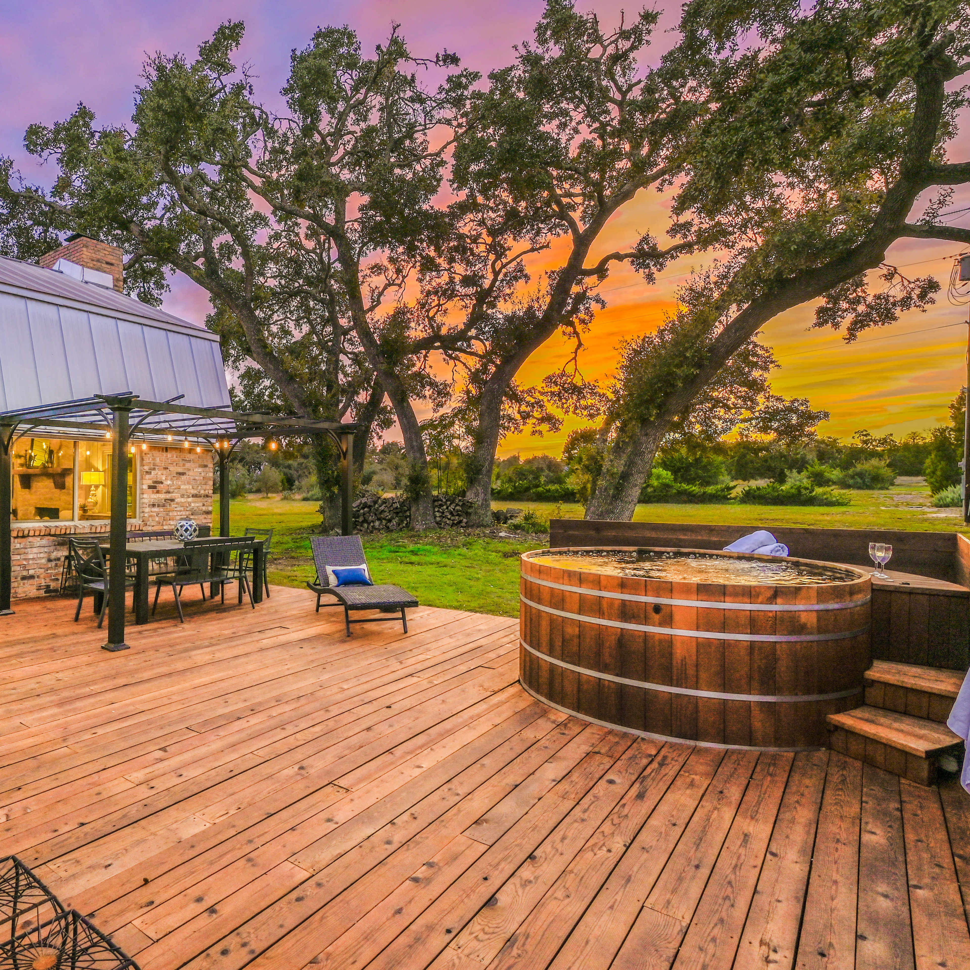 A wooden deck featuring a hot tub, outdoor seating, and large trees against a colorful sunset sky.