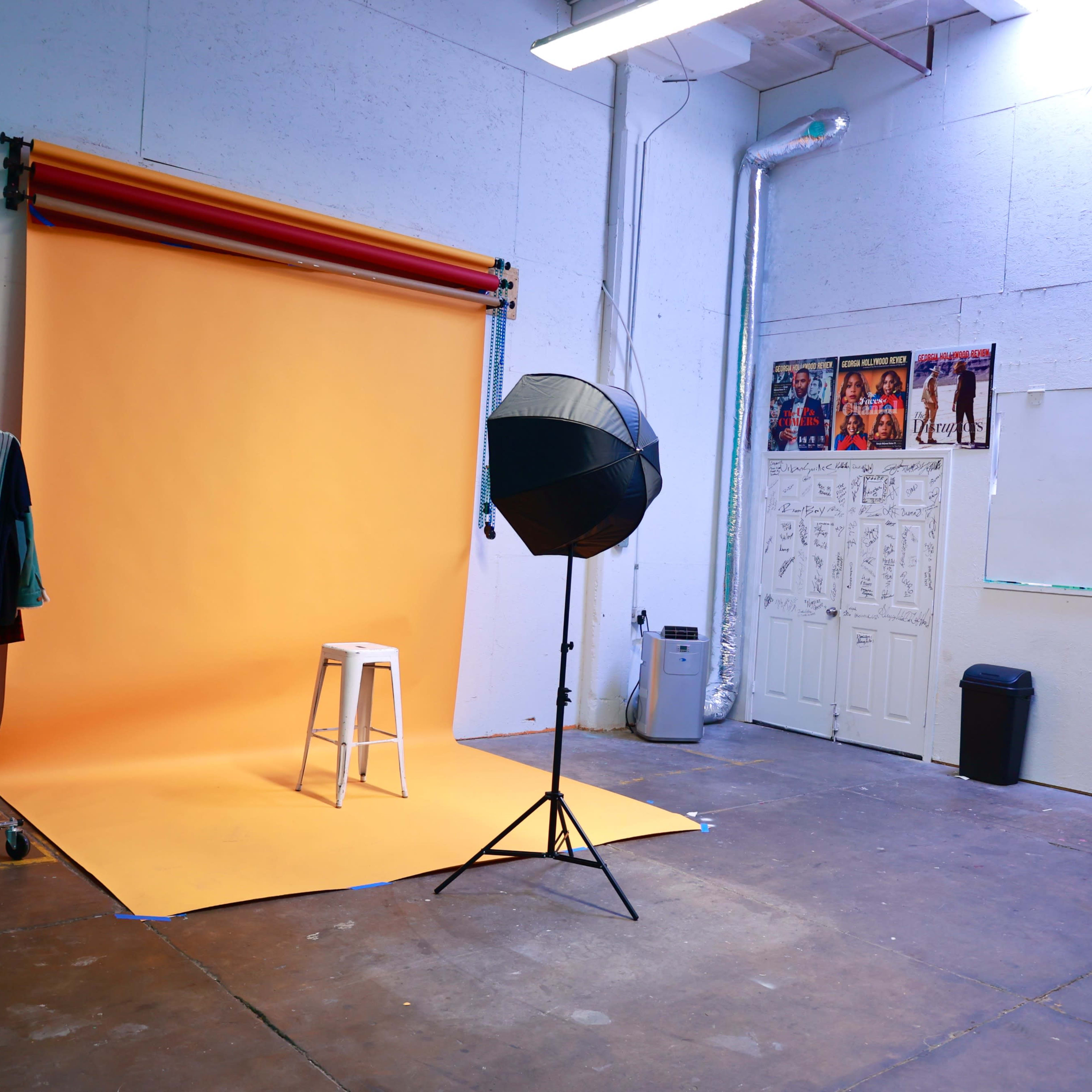A photography studio featuring an orange backdrop, a white stool, a softbox light, and a rack with clothing.