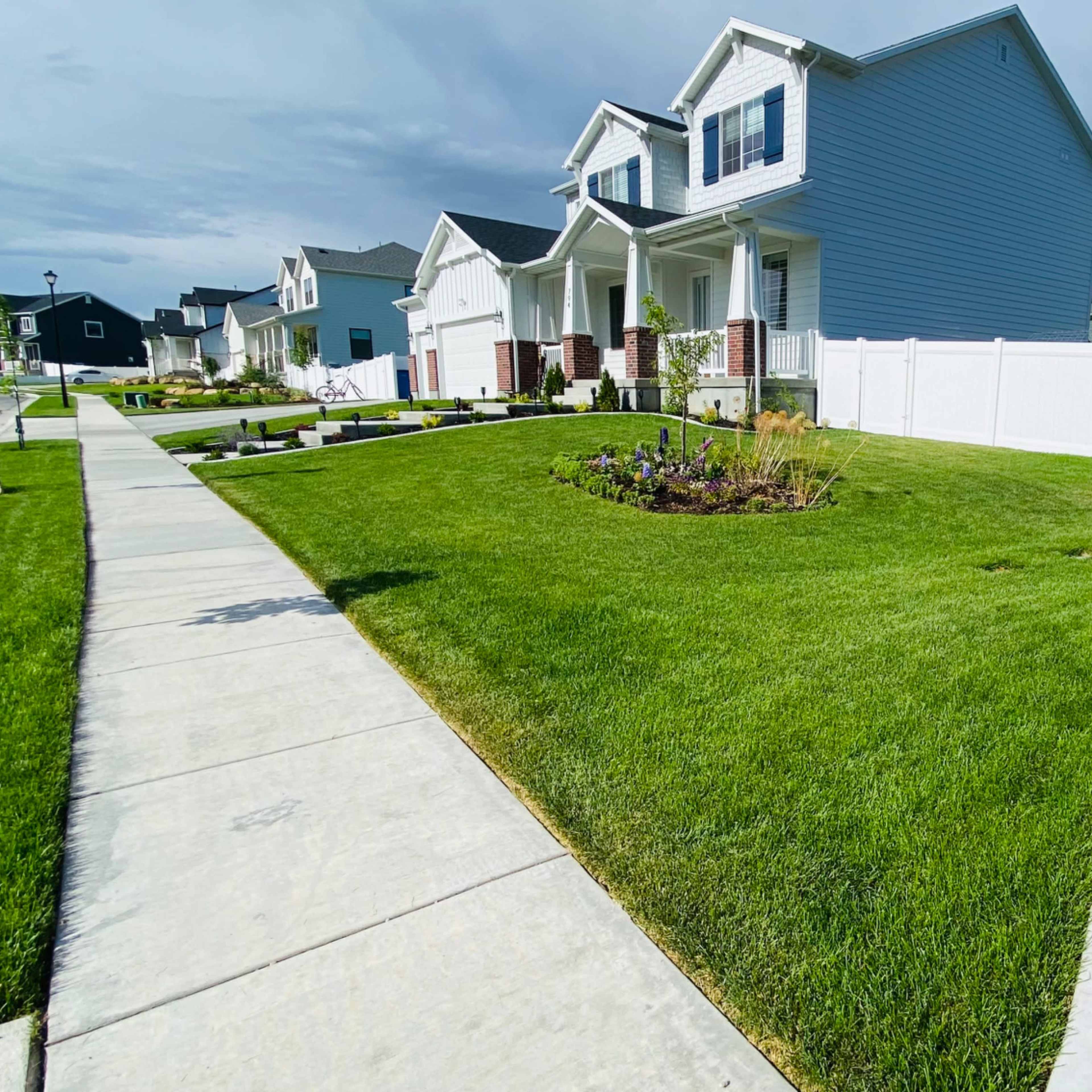 A well-maintained residential neighborhood features manicured lawns, flower beds, and concrete sidewalks.
