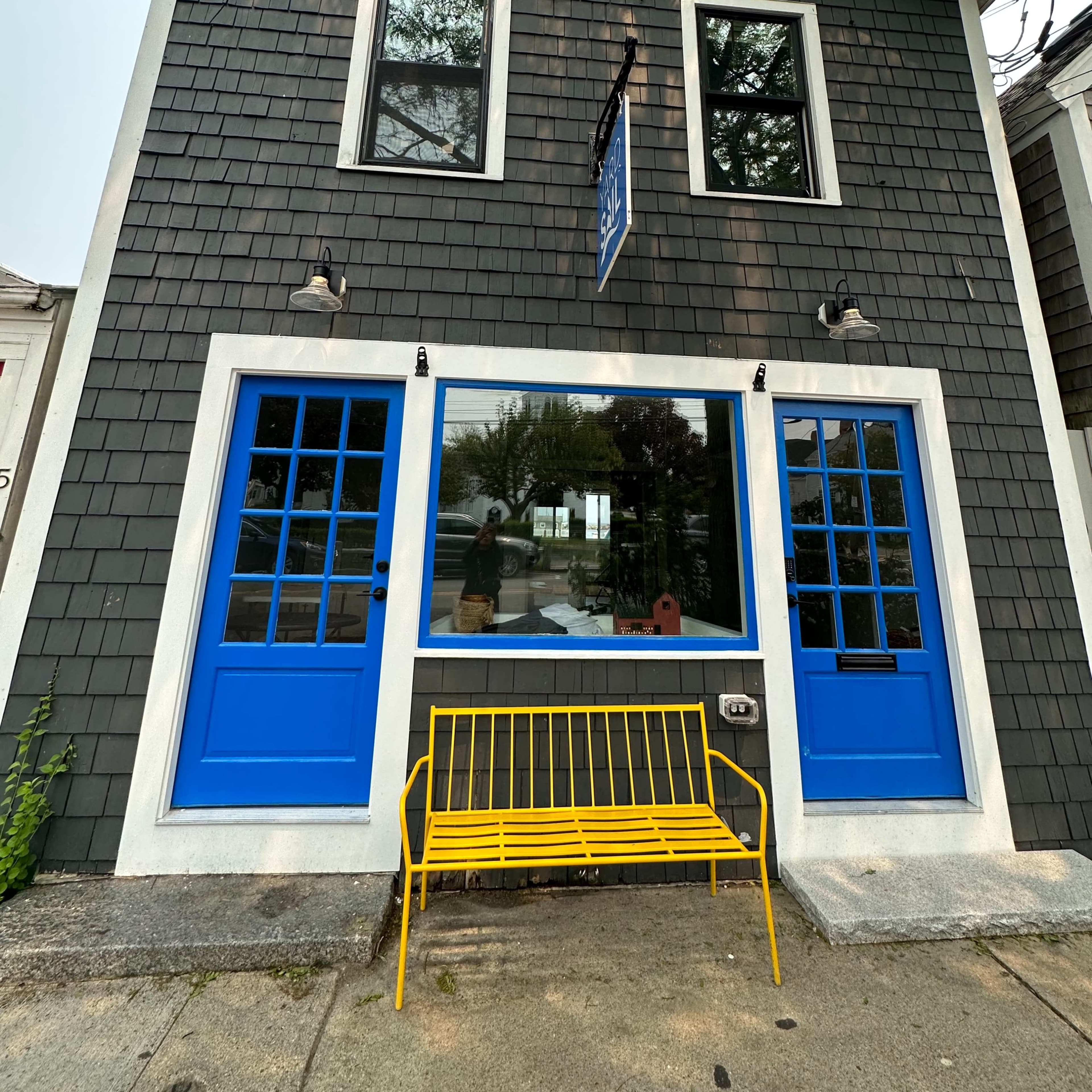 A two-story gray shingle building with bright blue double doors, a large central window, and a yellow bench in front.