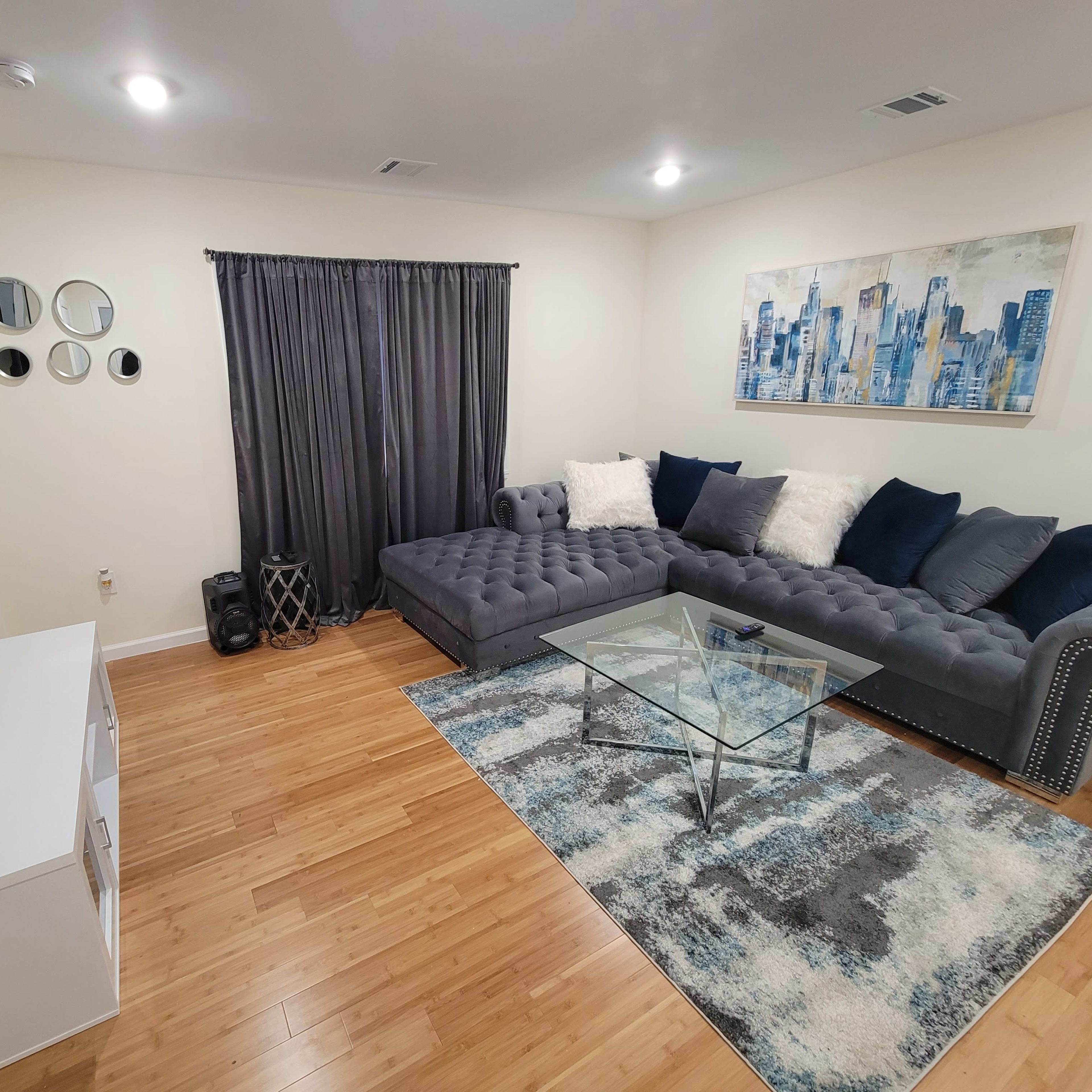 The room features a sectional sofa, a glass coffee table, a wall-mounted TV, a curtain, and decorative elements, all arranged on a hardwood floor.