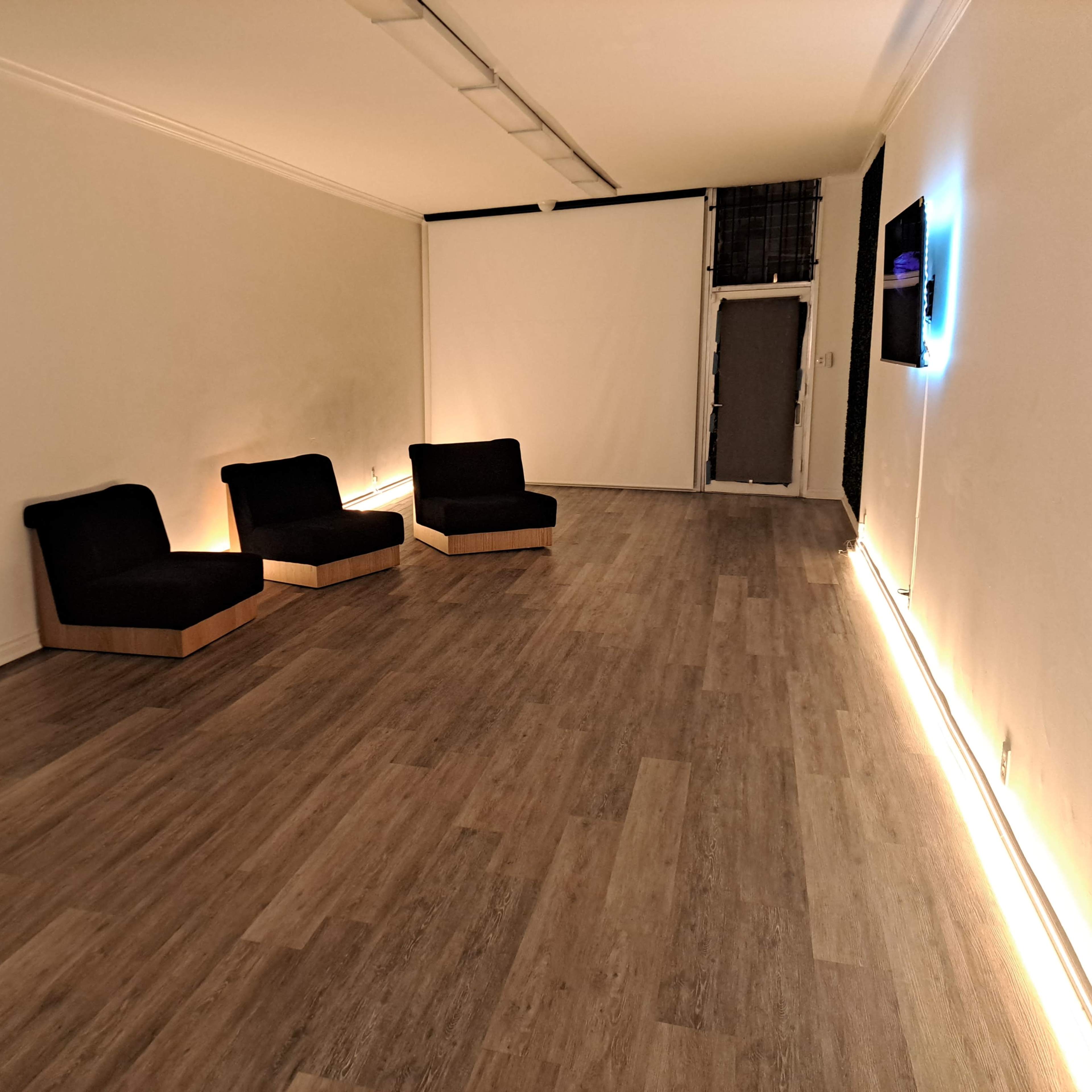A minimalist room with three black chairs positioned on a wooden floor, a large mirror on the left wall, and sparse lighting.
