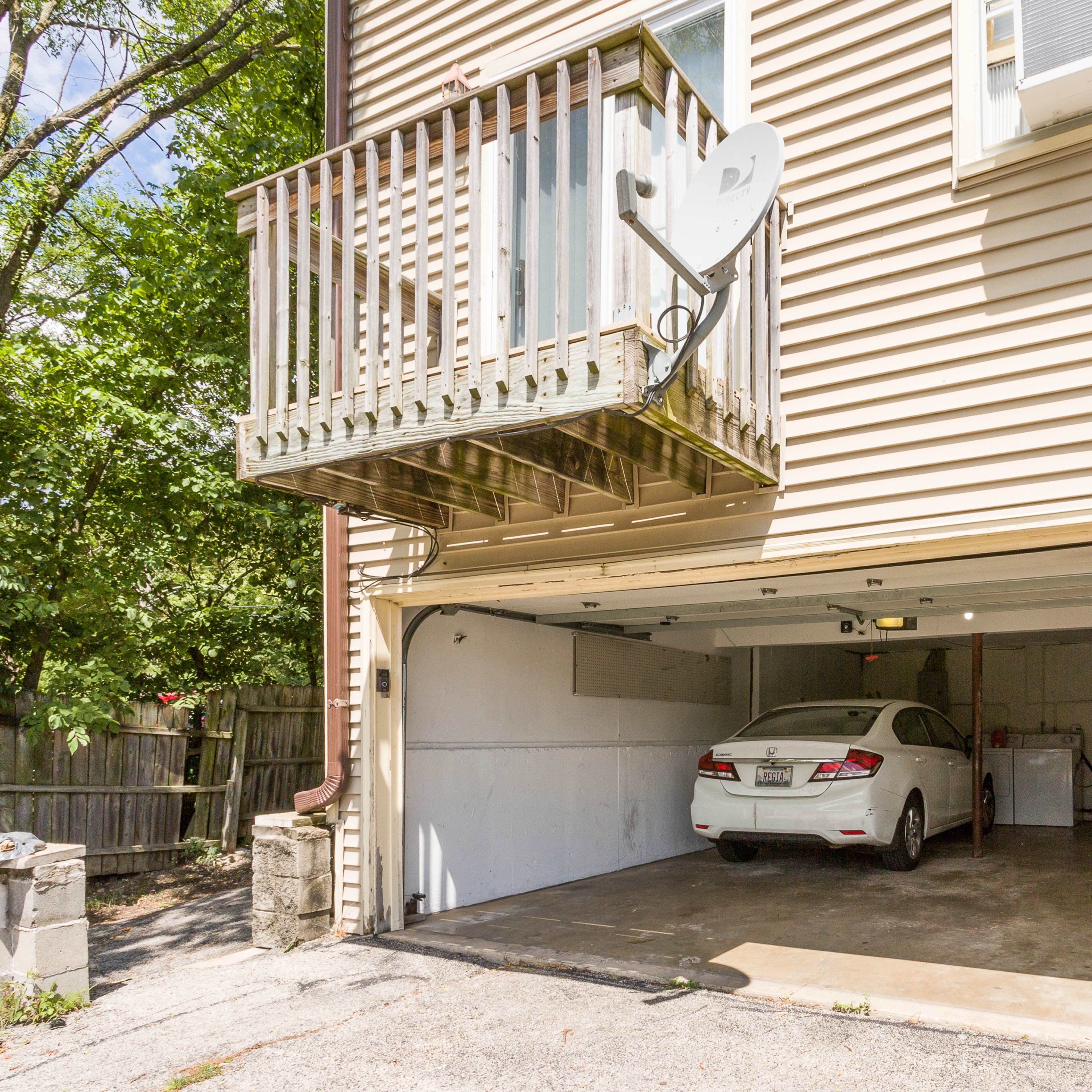 A white car is parked in a two-car garage beneath a balcony with a satellite dish attached to a beige house.