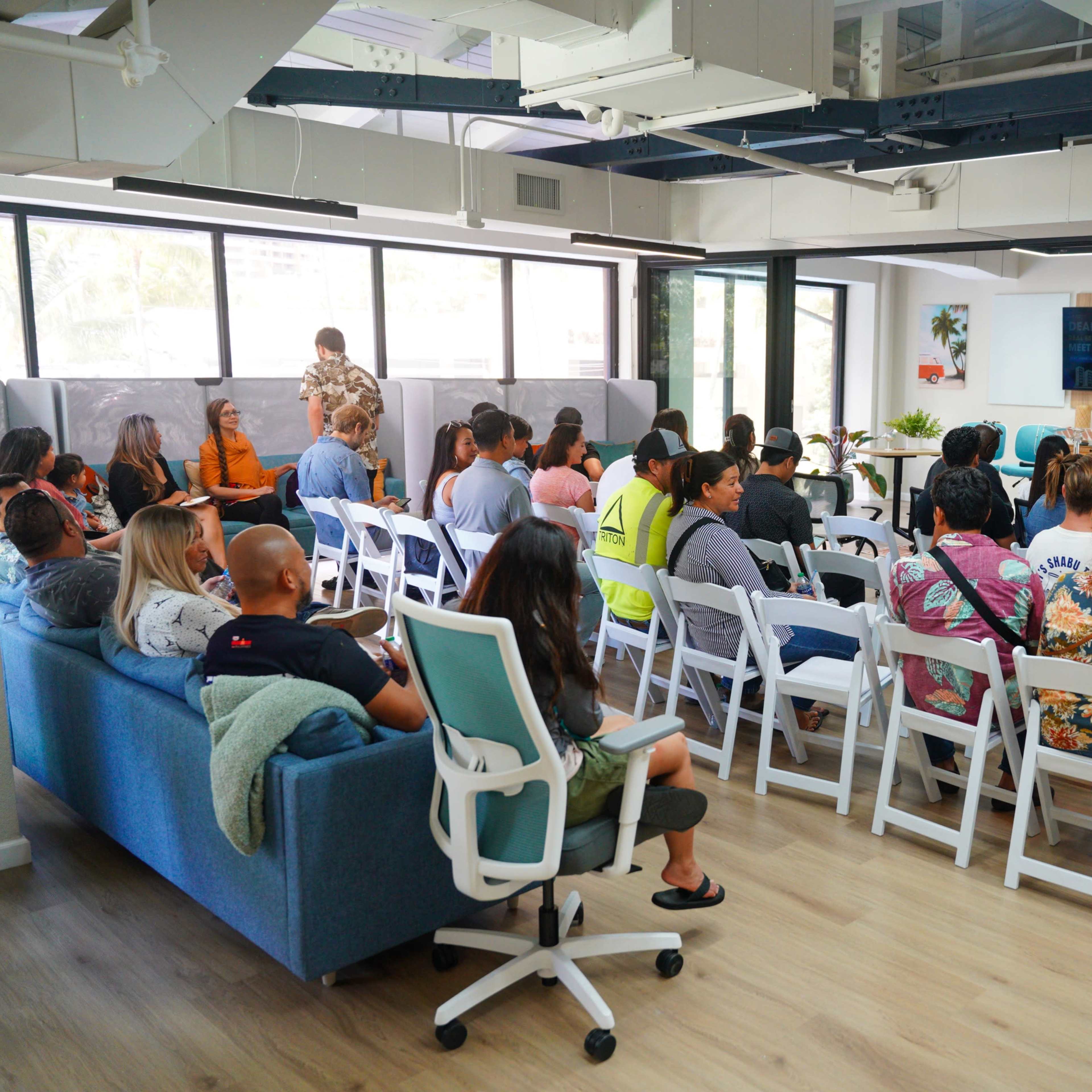 A group of people sits in rows of chairs facing a presenter in a modern office space with large windows and colorful decor.