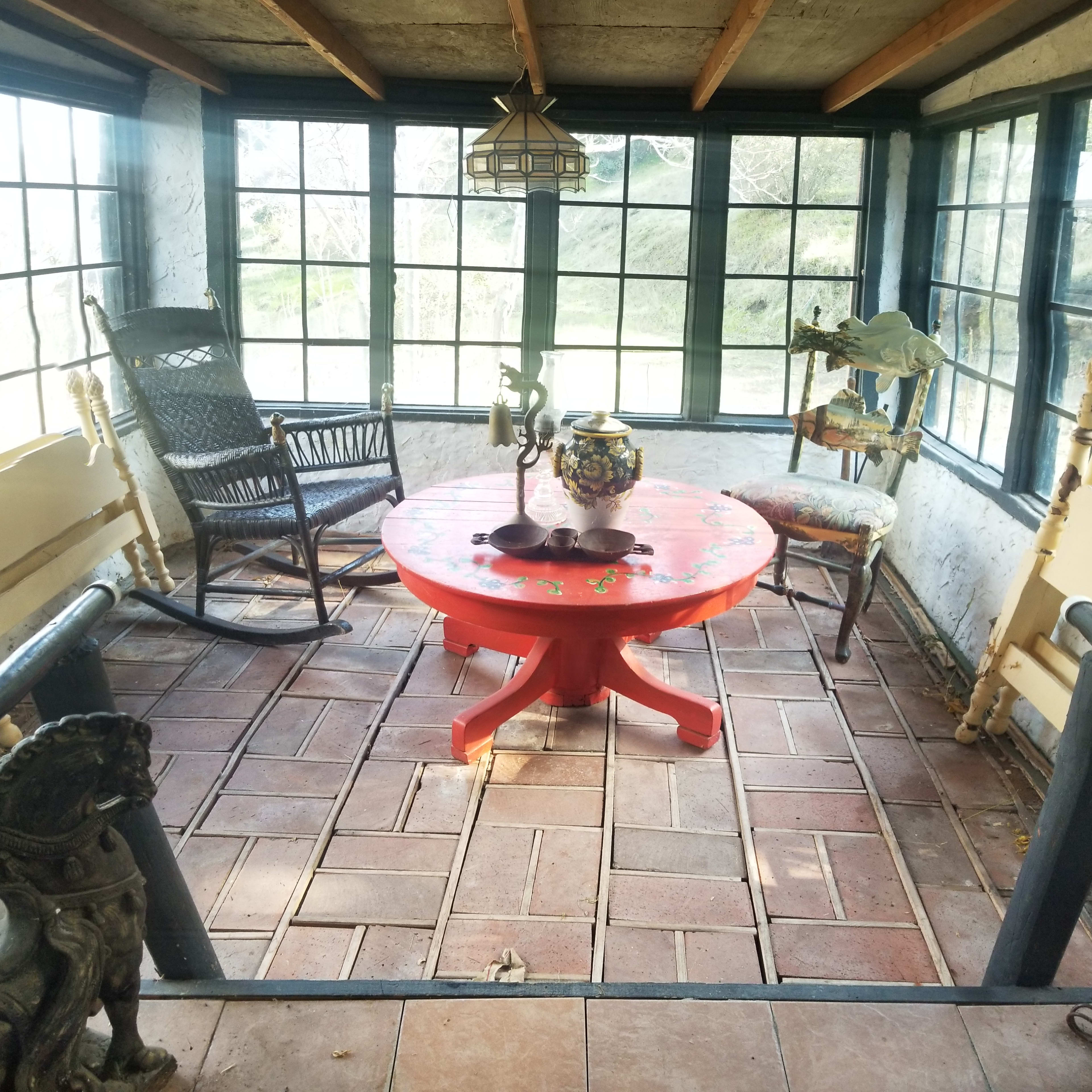 A sunlit room with large windows, featuring a red circular table, several chairs, and decorative items like a vase and a rocking chair.