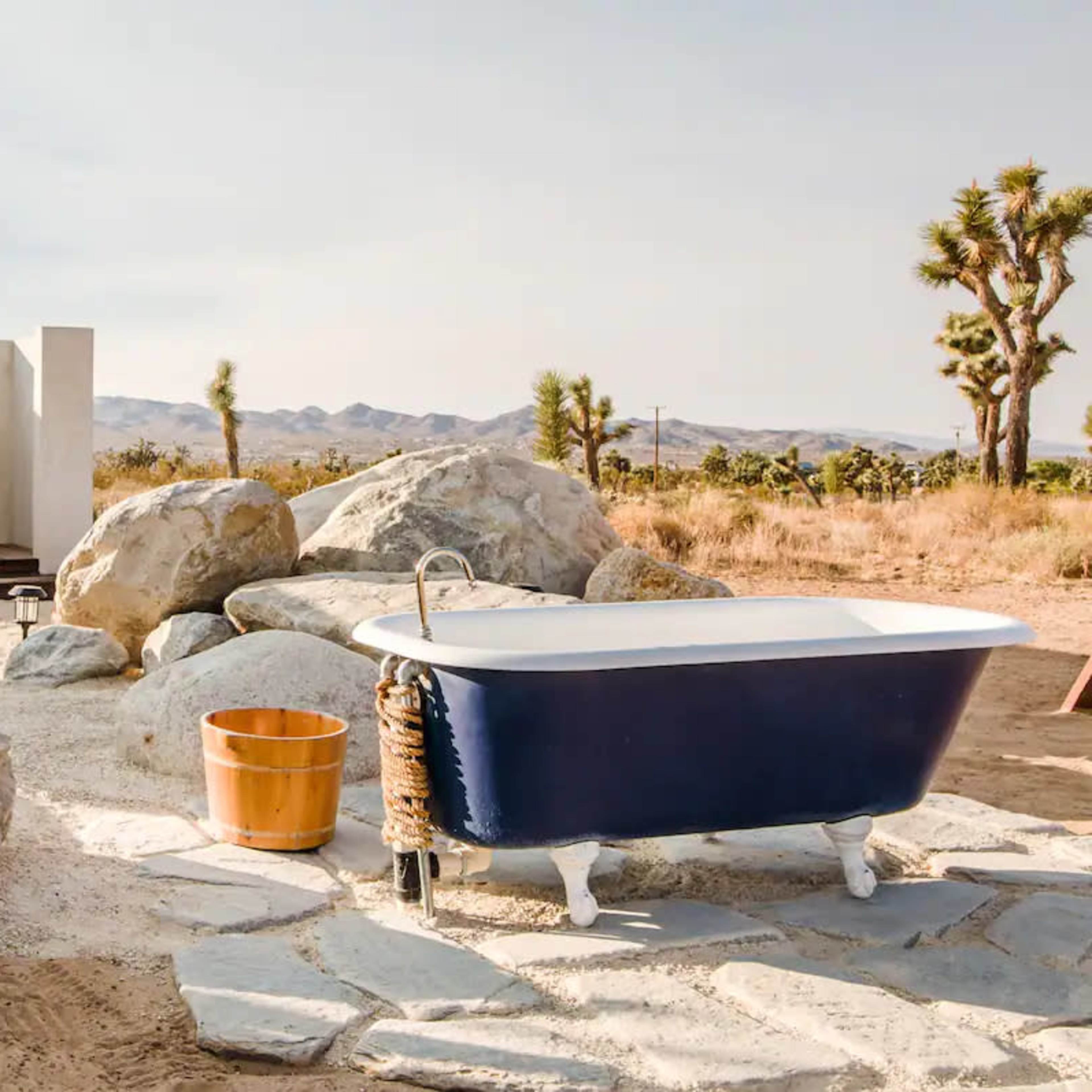 The scene features a freestanding bathtub next to large rocks and a small wooden structure in a desert landscape.