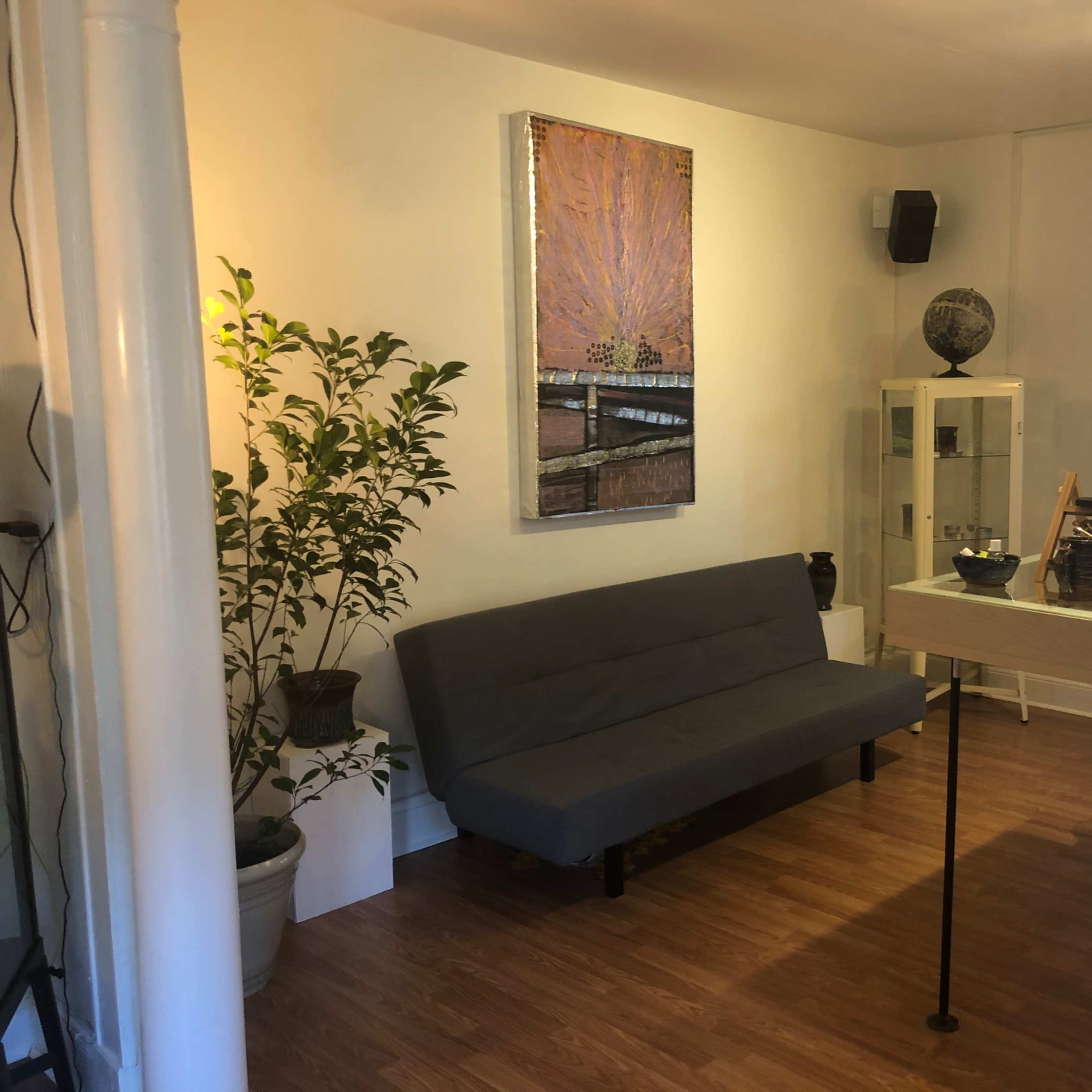 A cozy interior space featuring a gray sofa, a small table with items on it, and decorative plants, along with a large artwork on the wall.