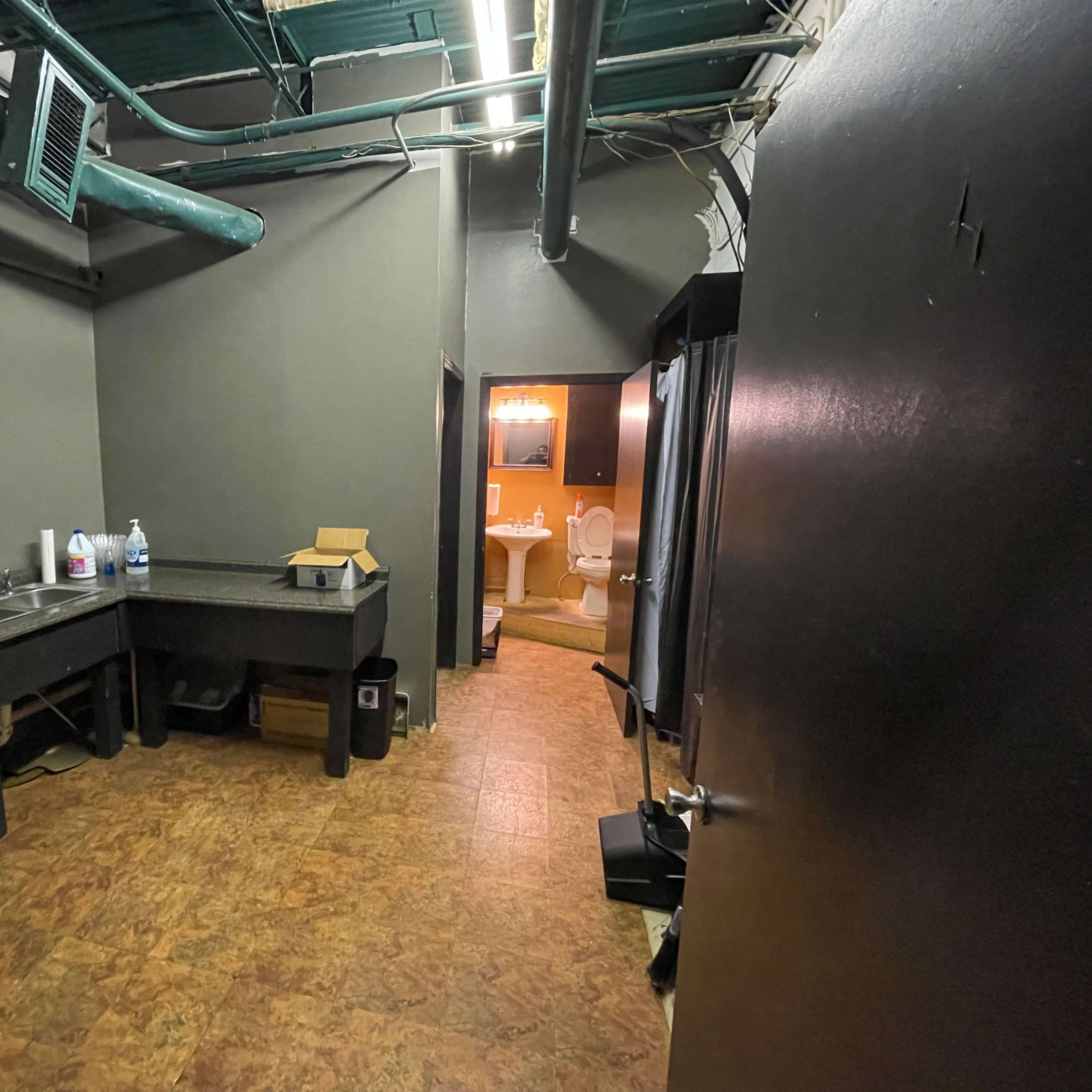 A dimly lit utility room with a refrigerator, sink, and a door leading to a bathroom in the background.