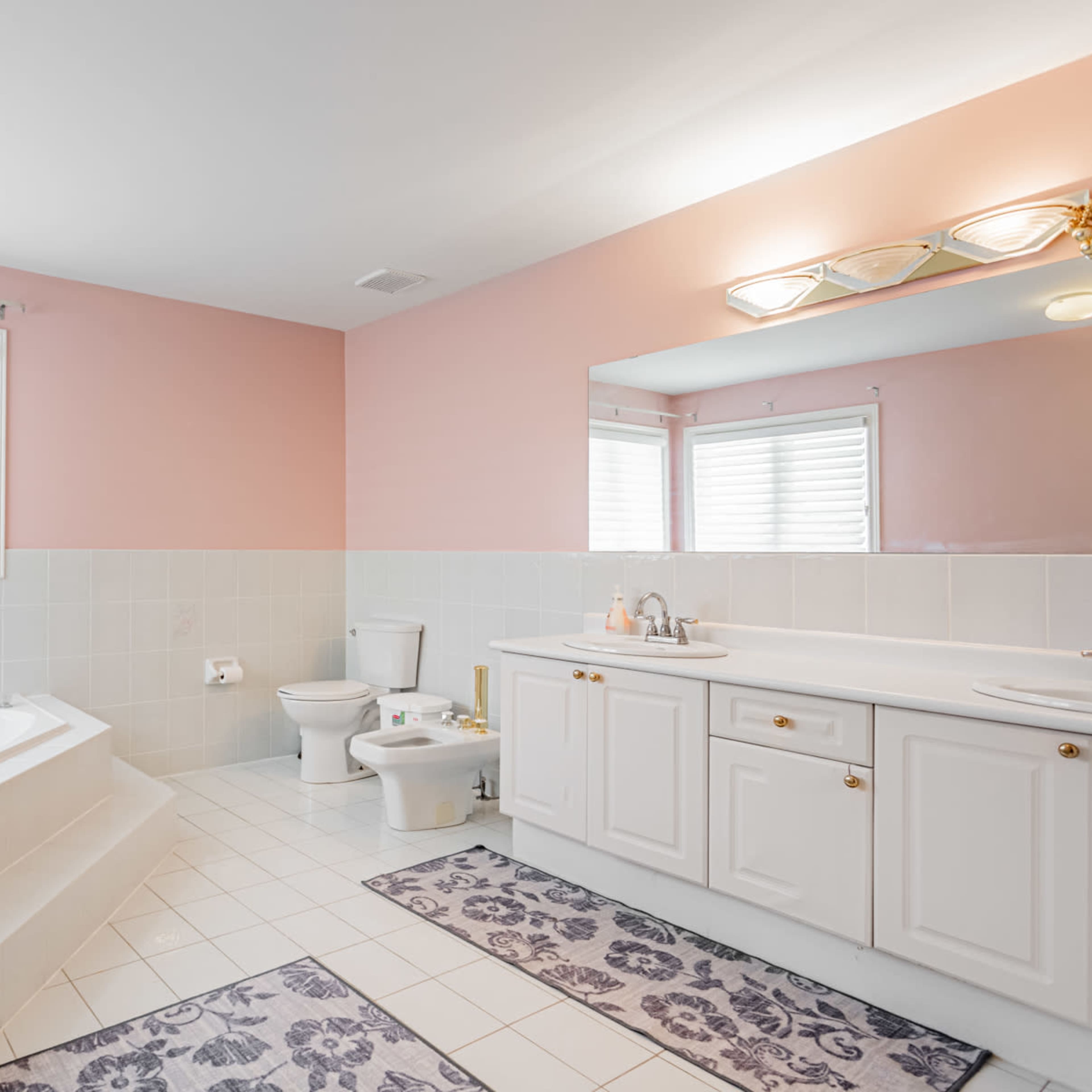A spacious bathroom features a corner bathtub, a toilet, and a double vanity with a large mirror, all set against light pink and white tiled walls.