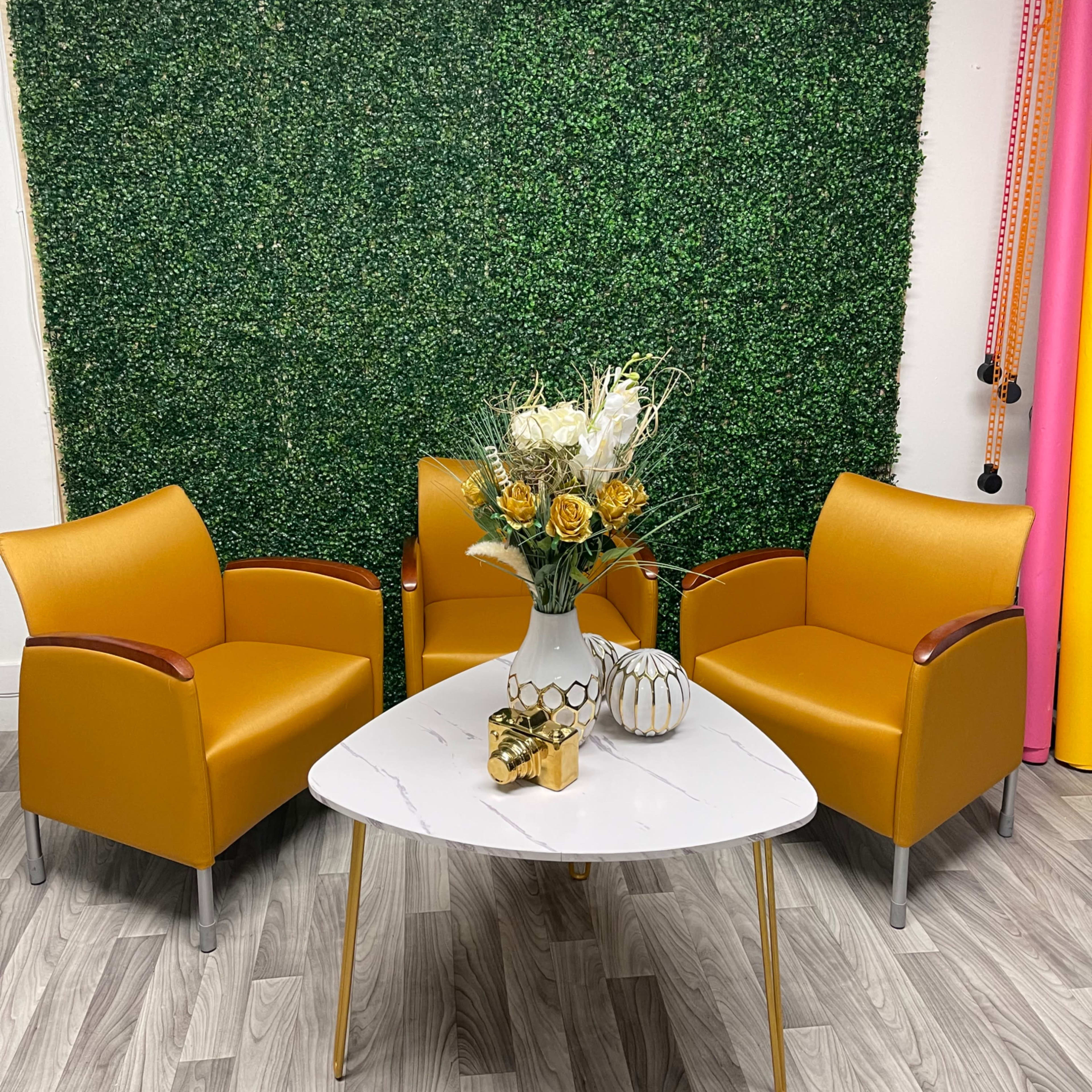 The scene features two mustard-colored chairs positioned around a white marble table with a floral arrangement and decorative items, set against a green hedge backdrop.