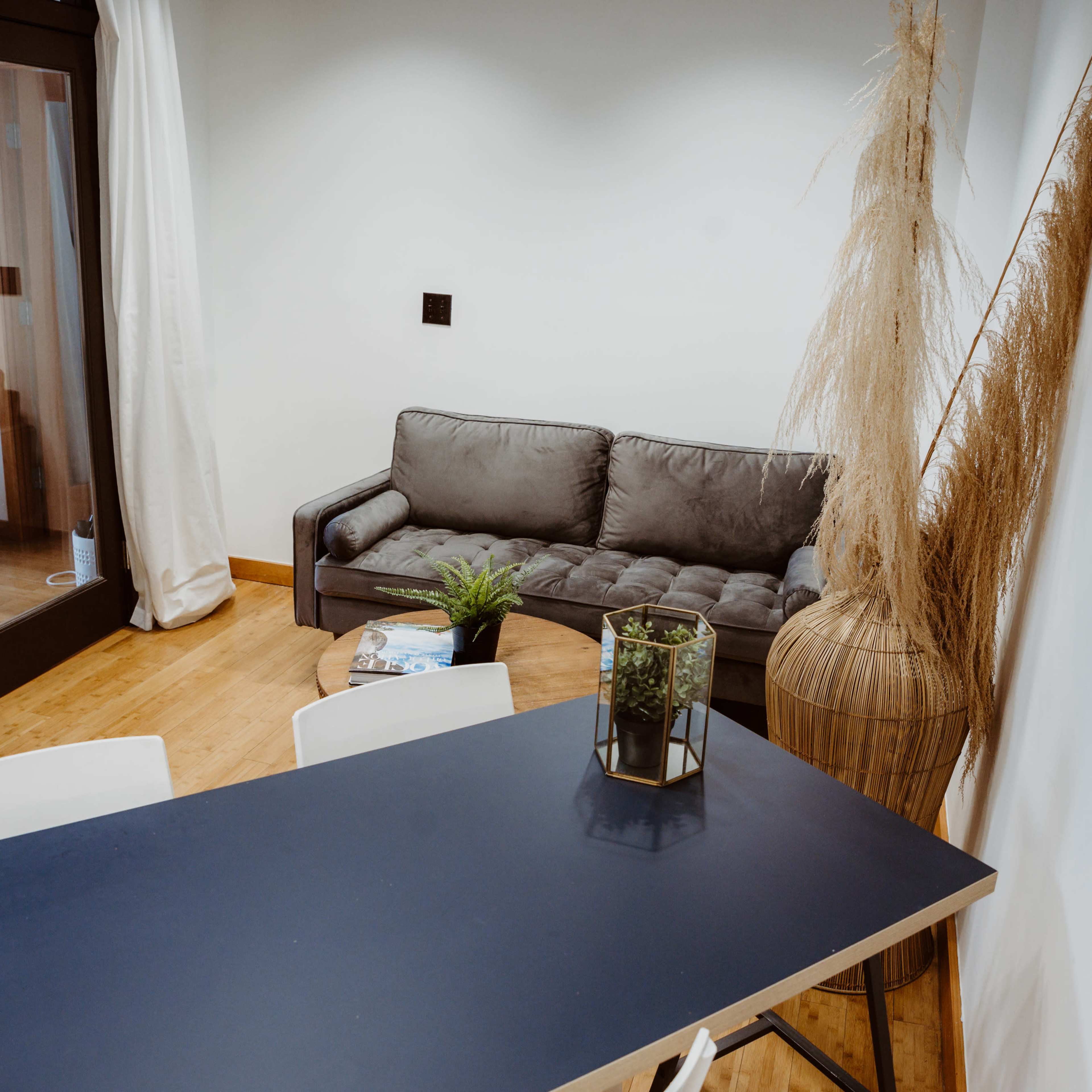 A modern living space features a dark table with white chairs, a gray sofa, and decorative elements like a vase and a plant.