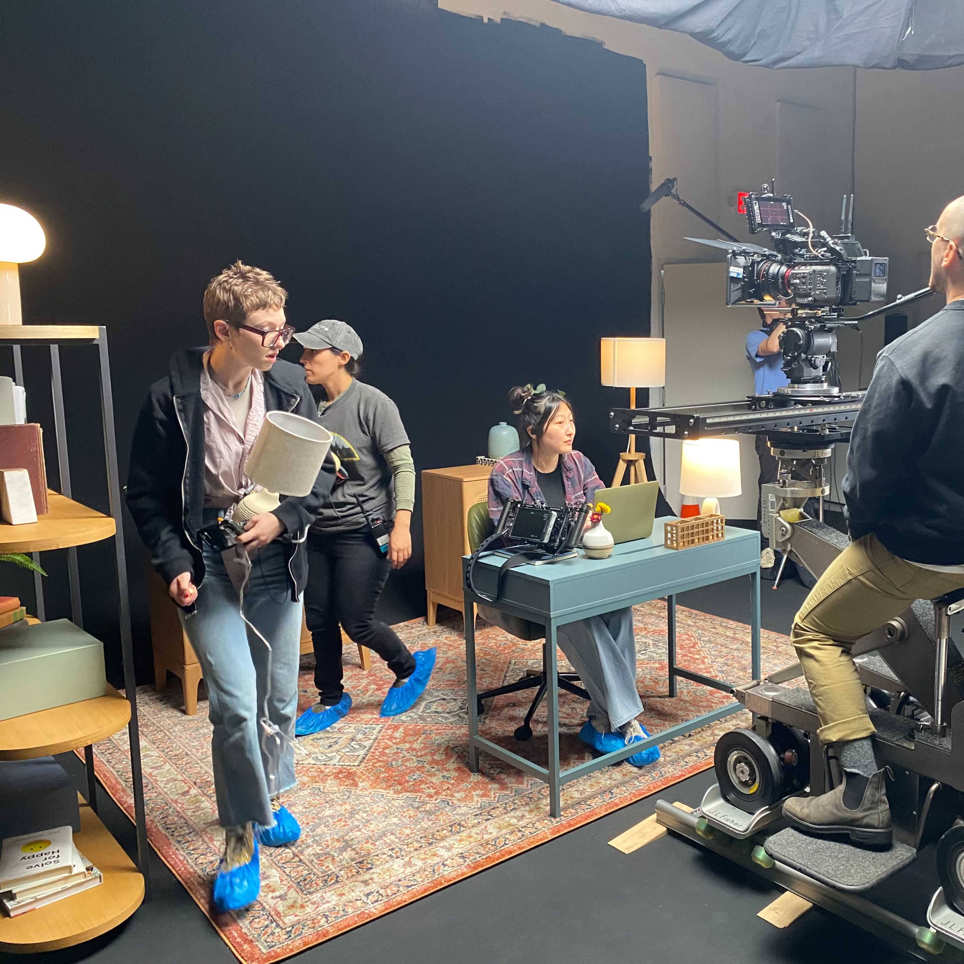 A film crew sets up a scene in a studio, featuring a desk with props and various lighting equipment.