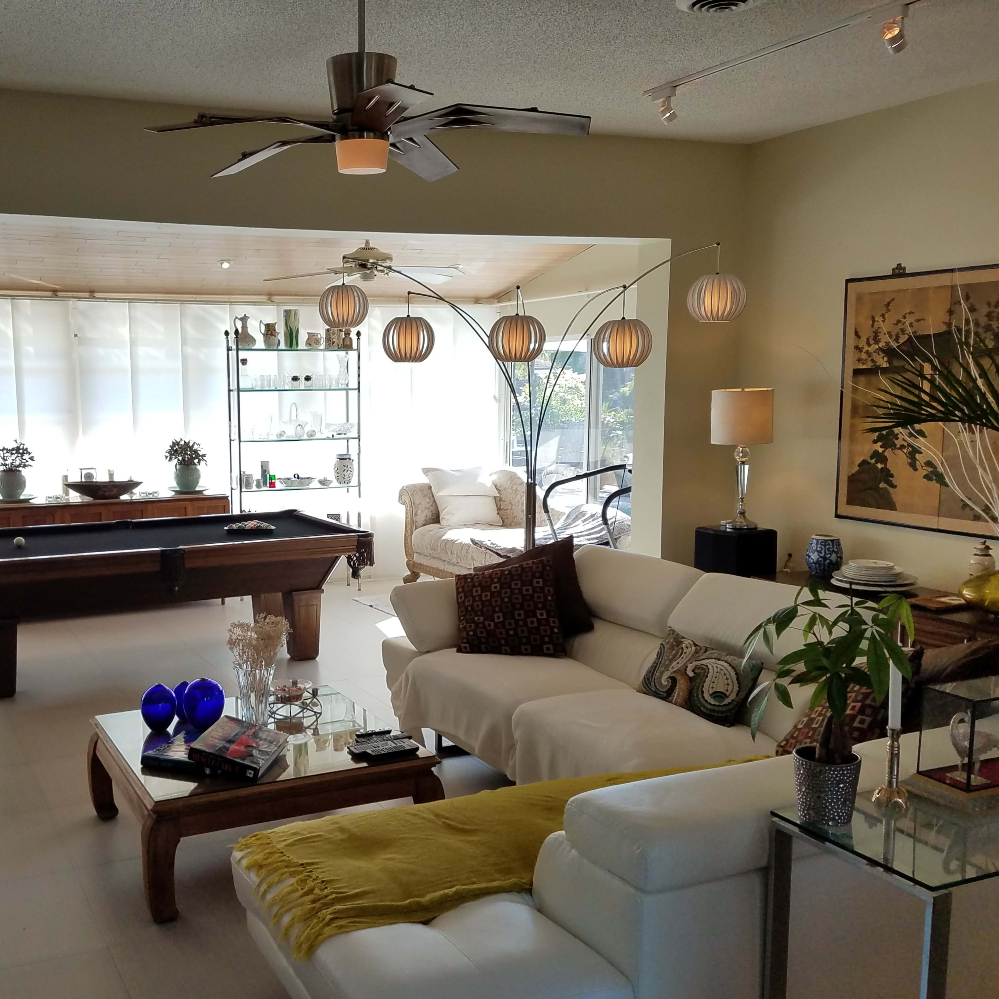 A spacious living area with a pool table, comfortable seating, decorative plants, and large windows allowing natural light to enter.