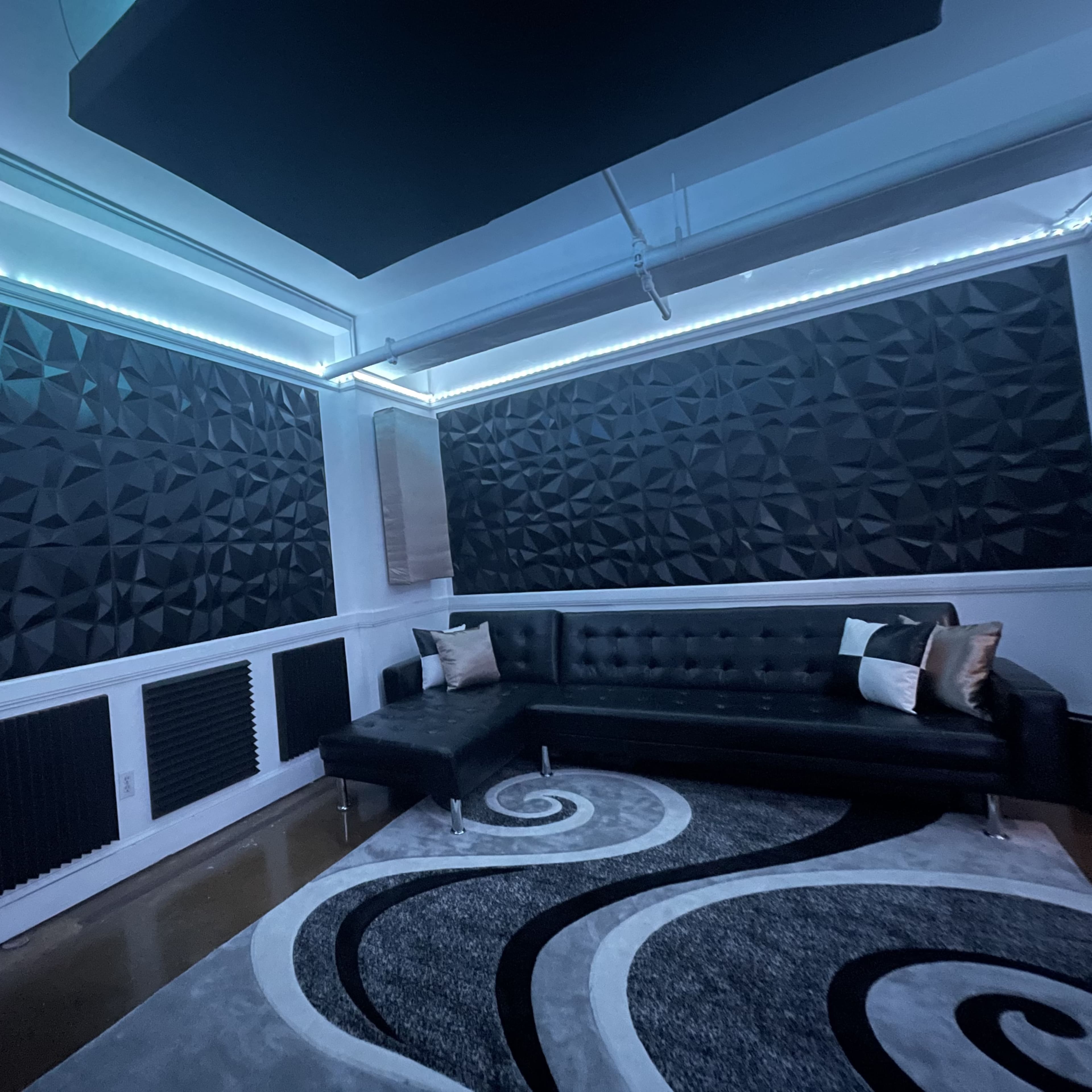 A modern room with a black leather sectional couch, angular acoustic wall panels, and a patterned area rug.