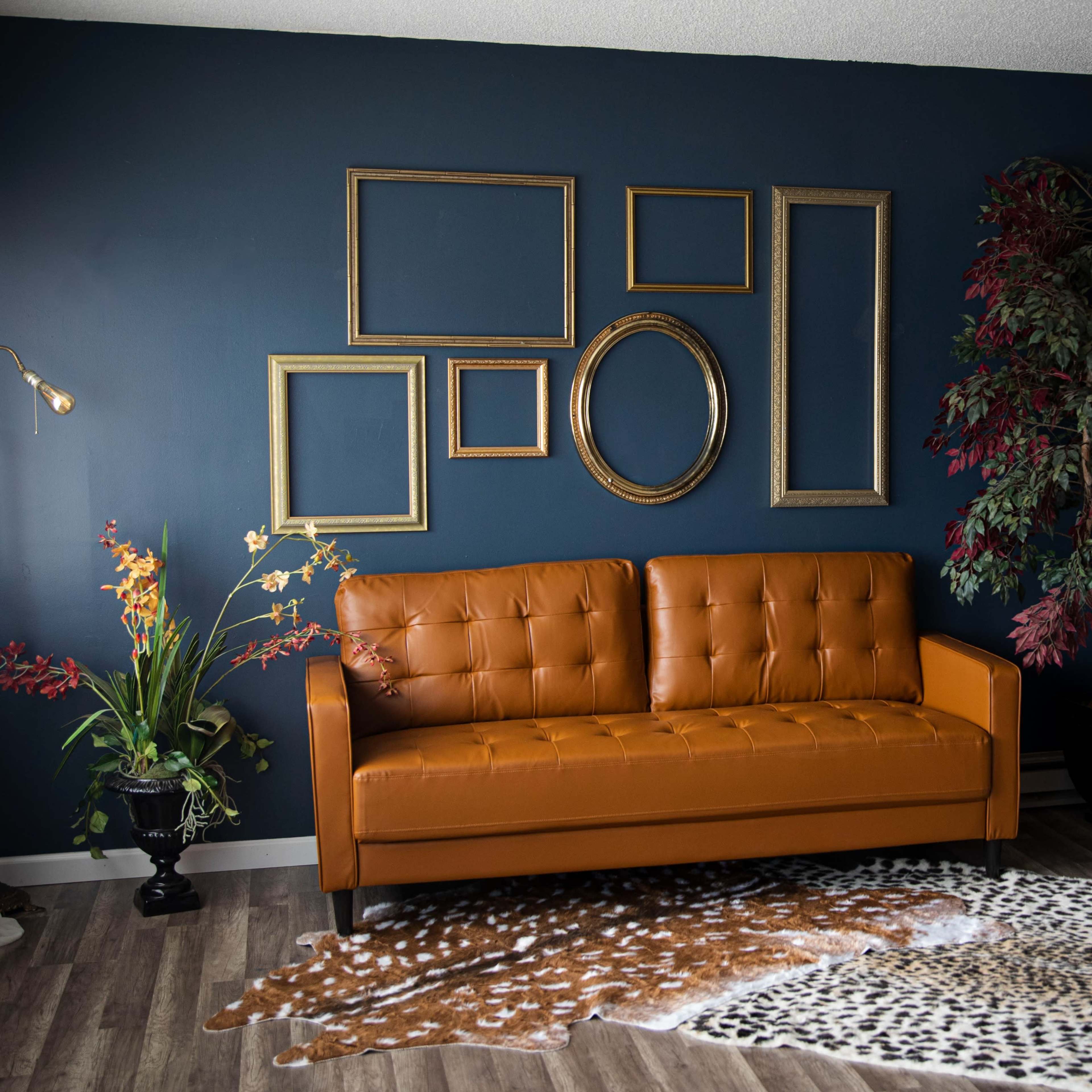 A modern living room features an orange leather sofa against a dark blue wall, adorned with empty picture frames and a decorative tree, alongside a patterned rug on the floor.
