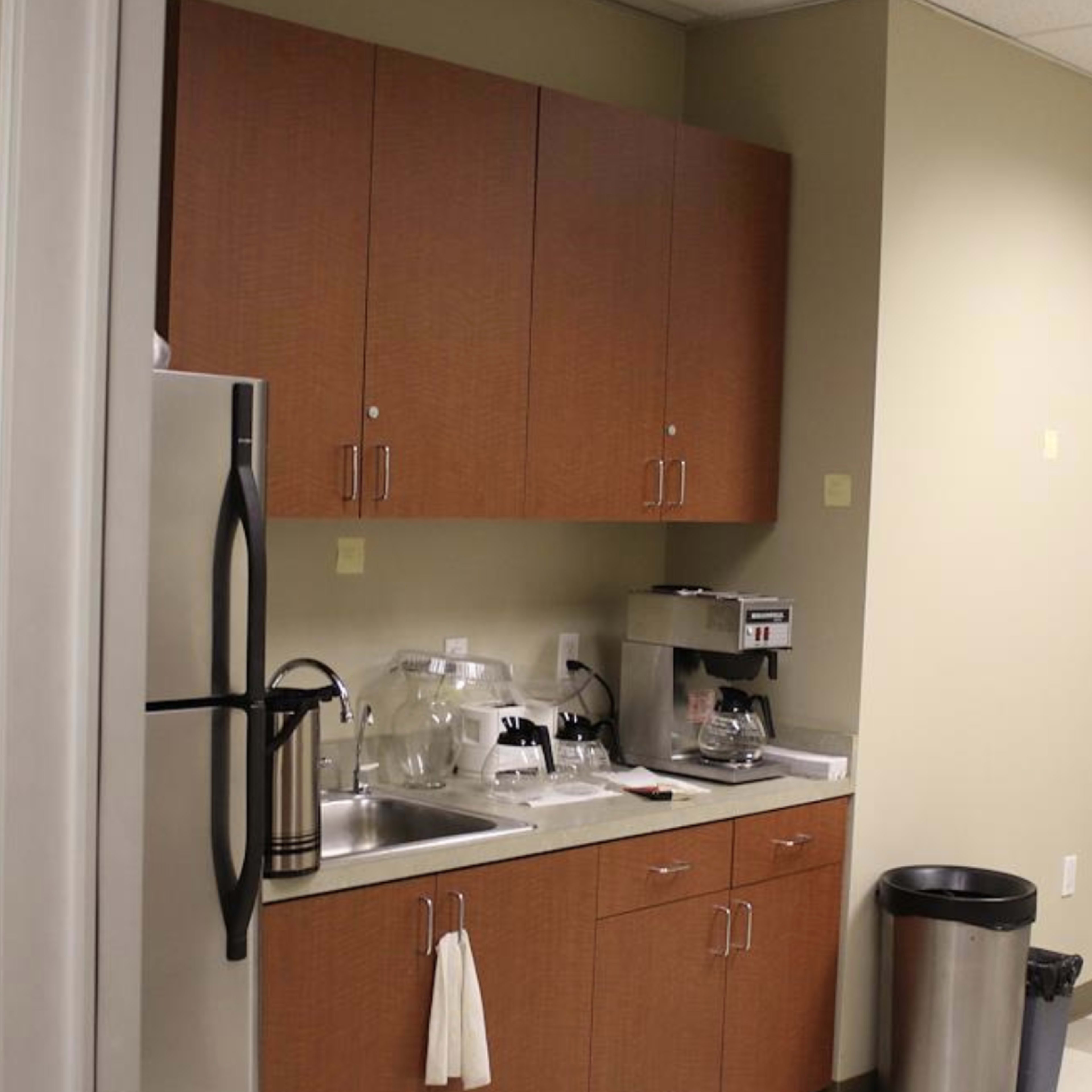 A small kitchenette area with a stainless steel refrigerator, coffee maker, sink, and cabinetry against a neutral-colored wall.