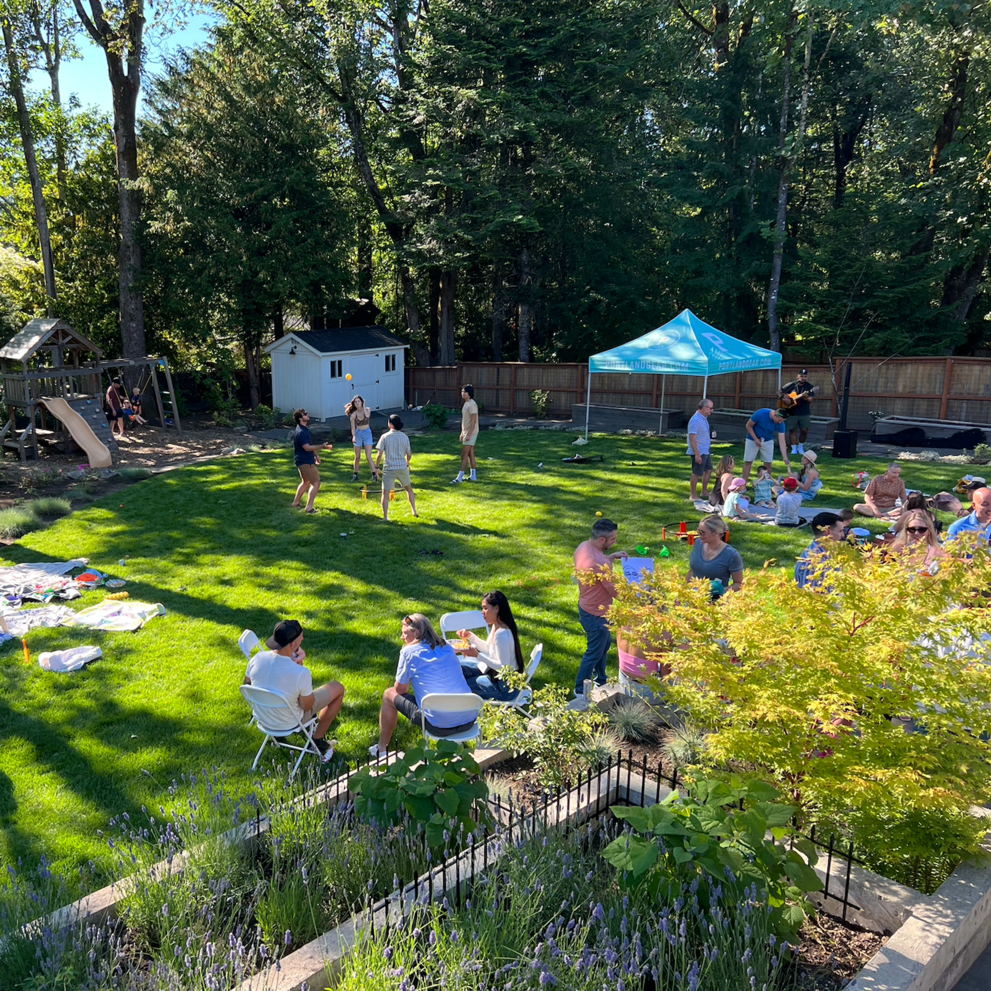 A group of people gathers on a grassy backyard for a picnic, with children playing nearby and a blue tent set up for shade.