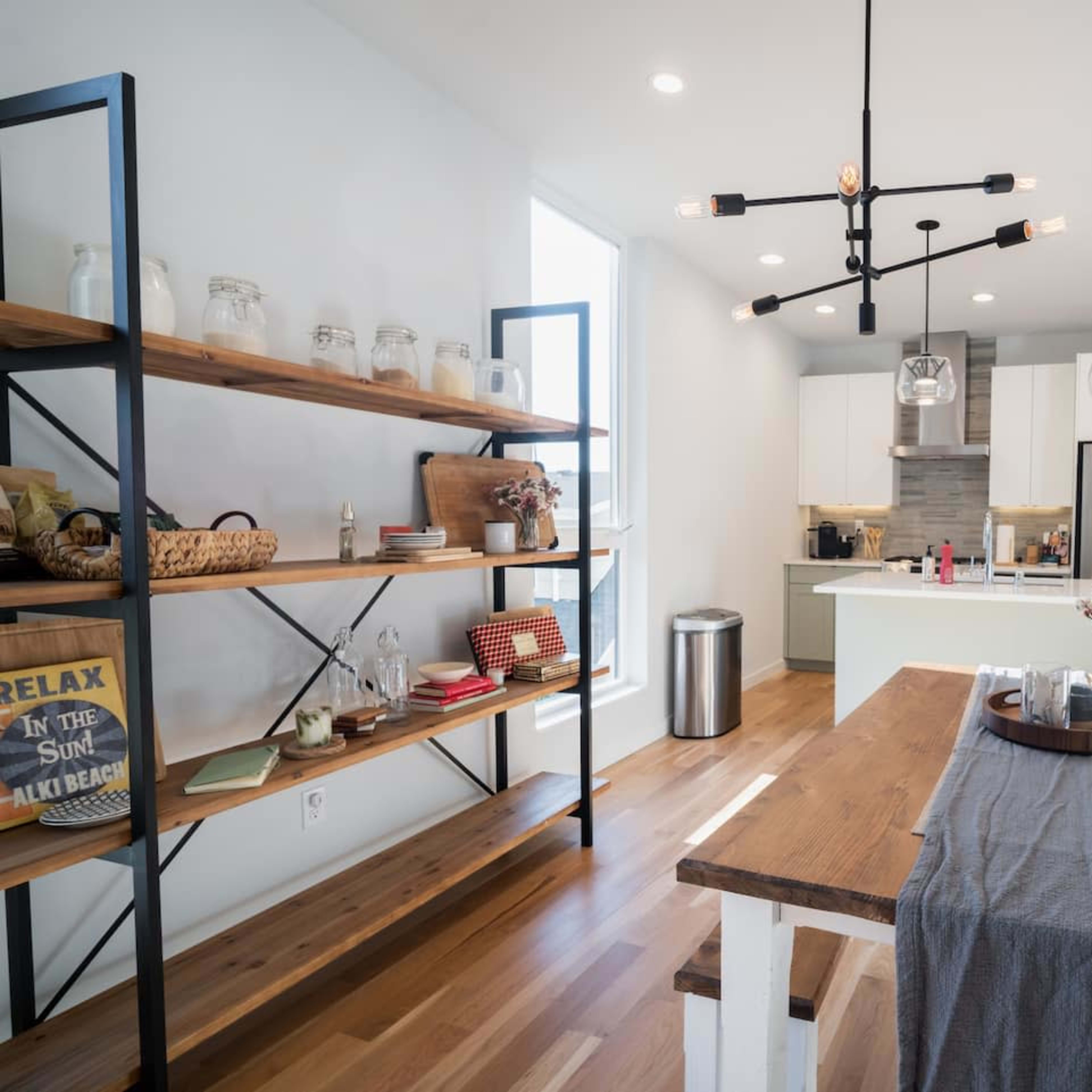 A modern kitchen with wooden shelves, a dining table, and stainless steel appliances, featuring large windows that provide ample natural light.