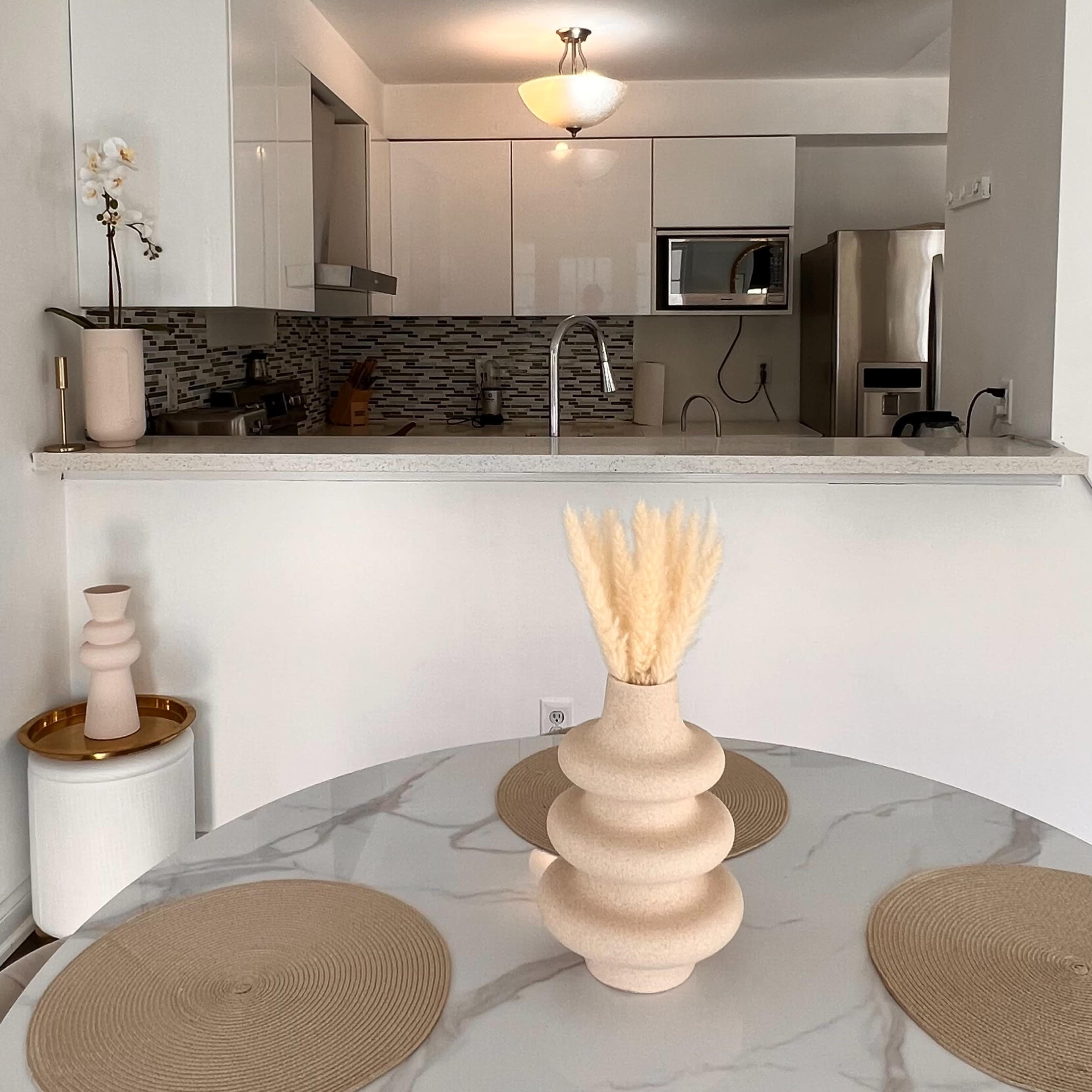 A modern kitchen and dining area feature a round table set with placemats and a decorative vase, with the kitchen visible in the background.