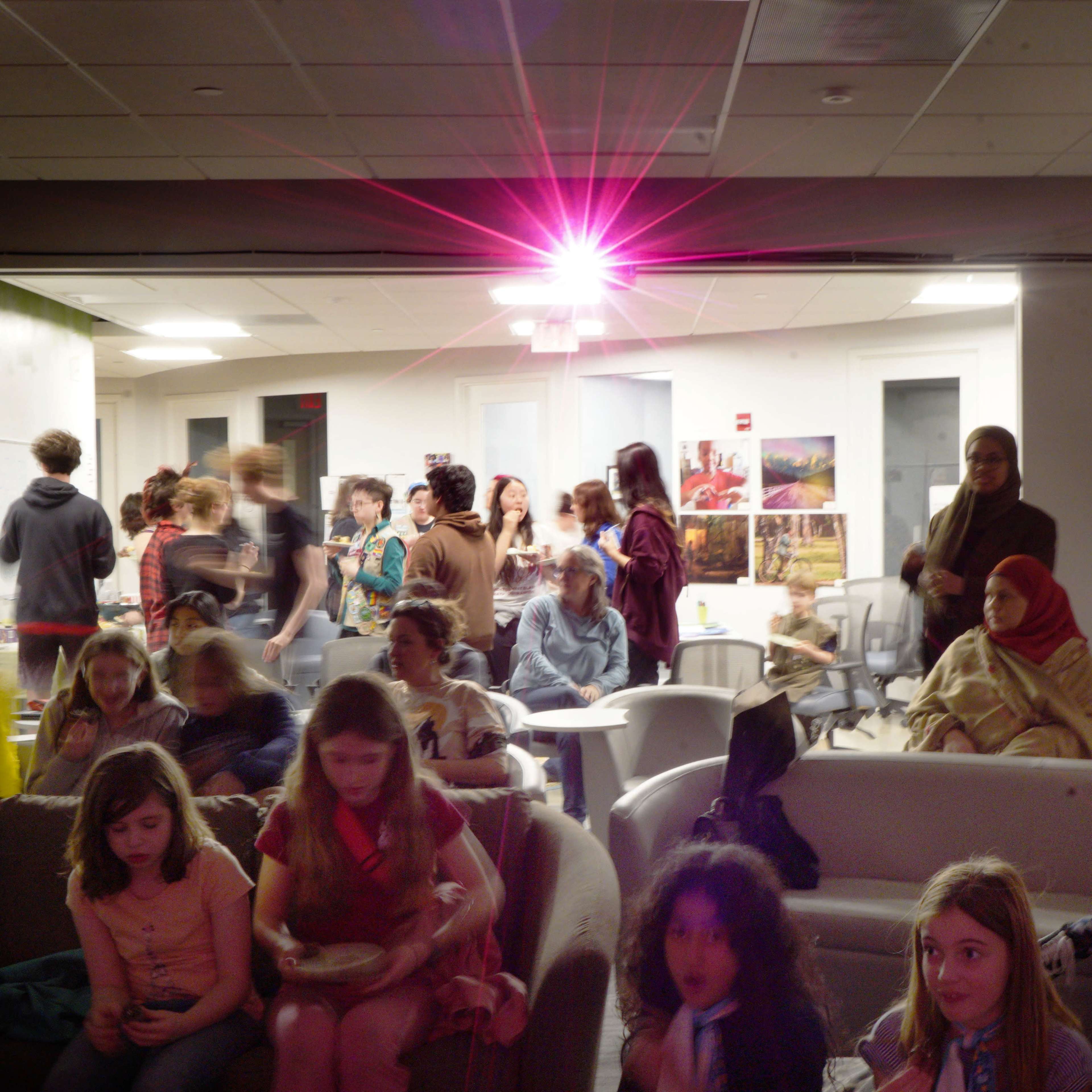 A group of people gather in a room with a large light source, while some sit on a couch and others stand or converse in the background.