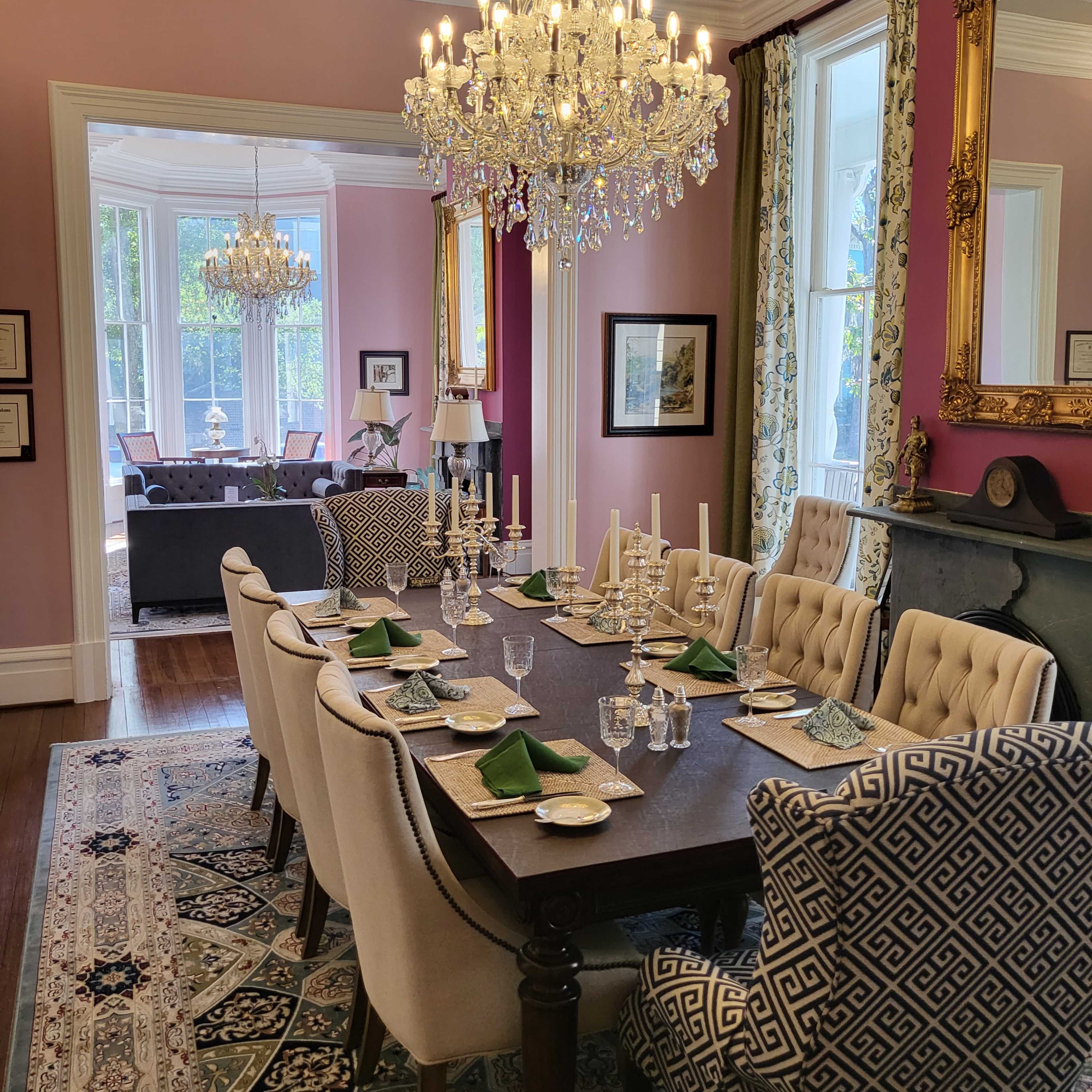 A long dining table is set with elegant dishware, candles, and green napkins under a large chandelier in a pink-walled room with windows overlooking a garden.