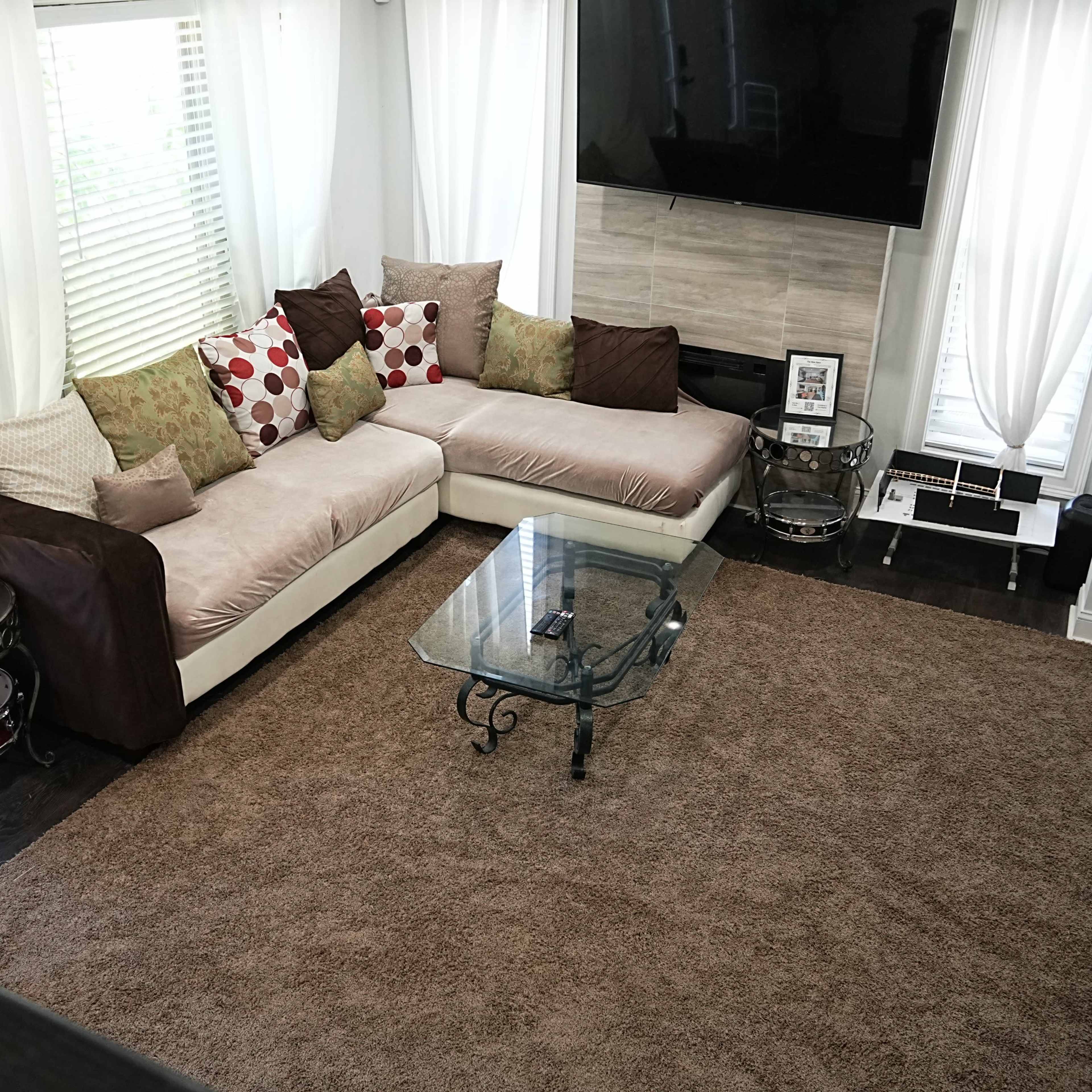 A living room with a sectional sofa, a glass coffee table, and a large area rug, all arranged around a wall-mounted television and large windows with sheer curtains.