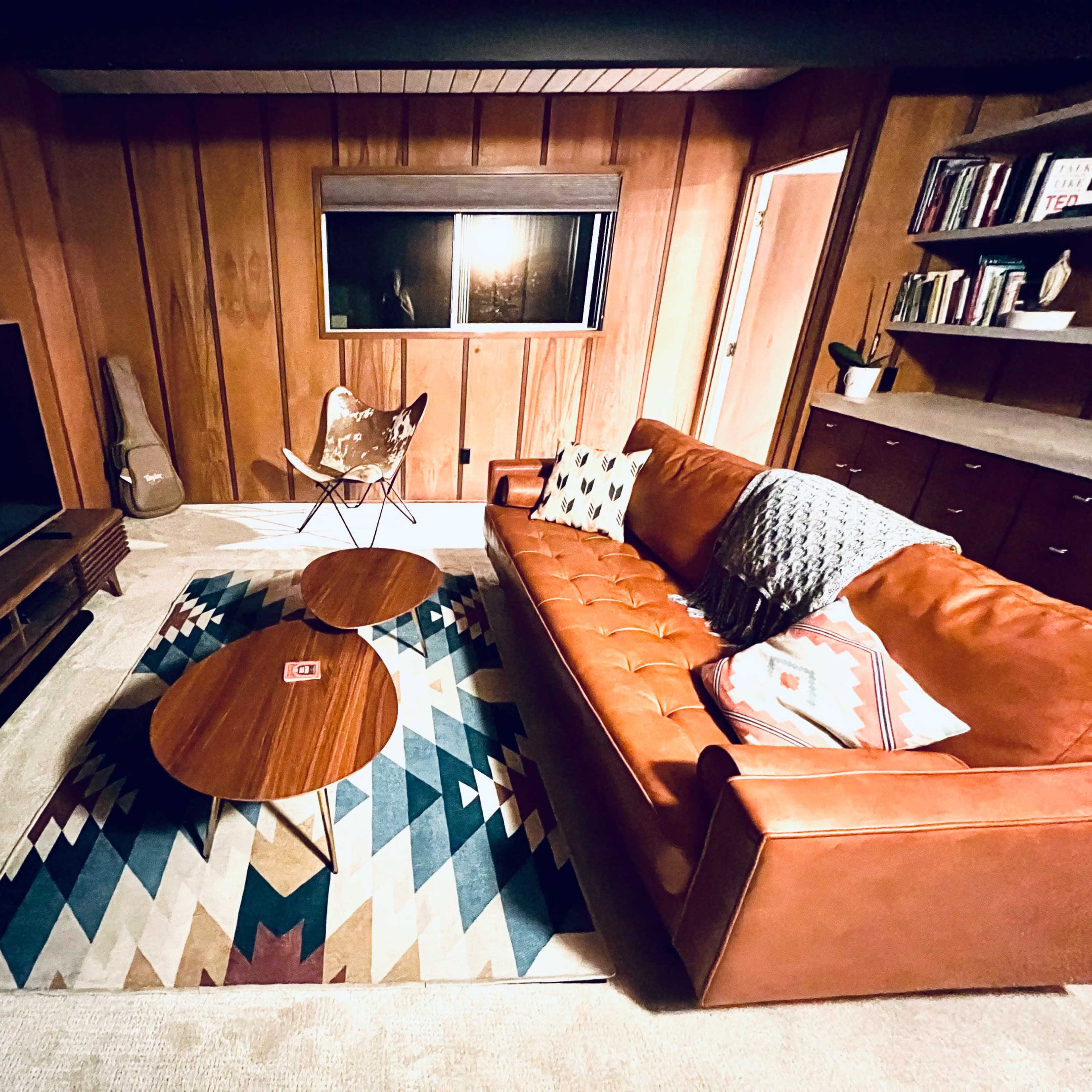 A cozy living room with a brown leather sofa, two wooden coffee tables, a patterned rug, and a large television against wooden-paneled walls.