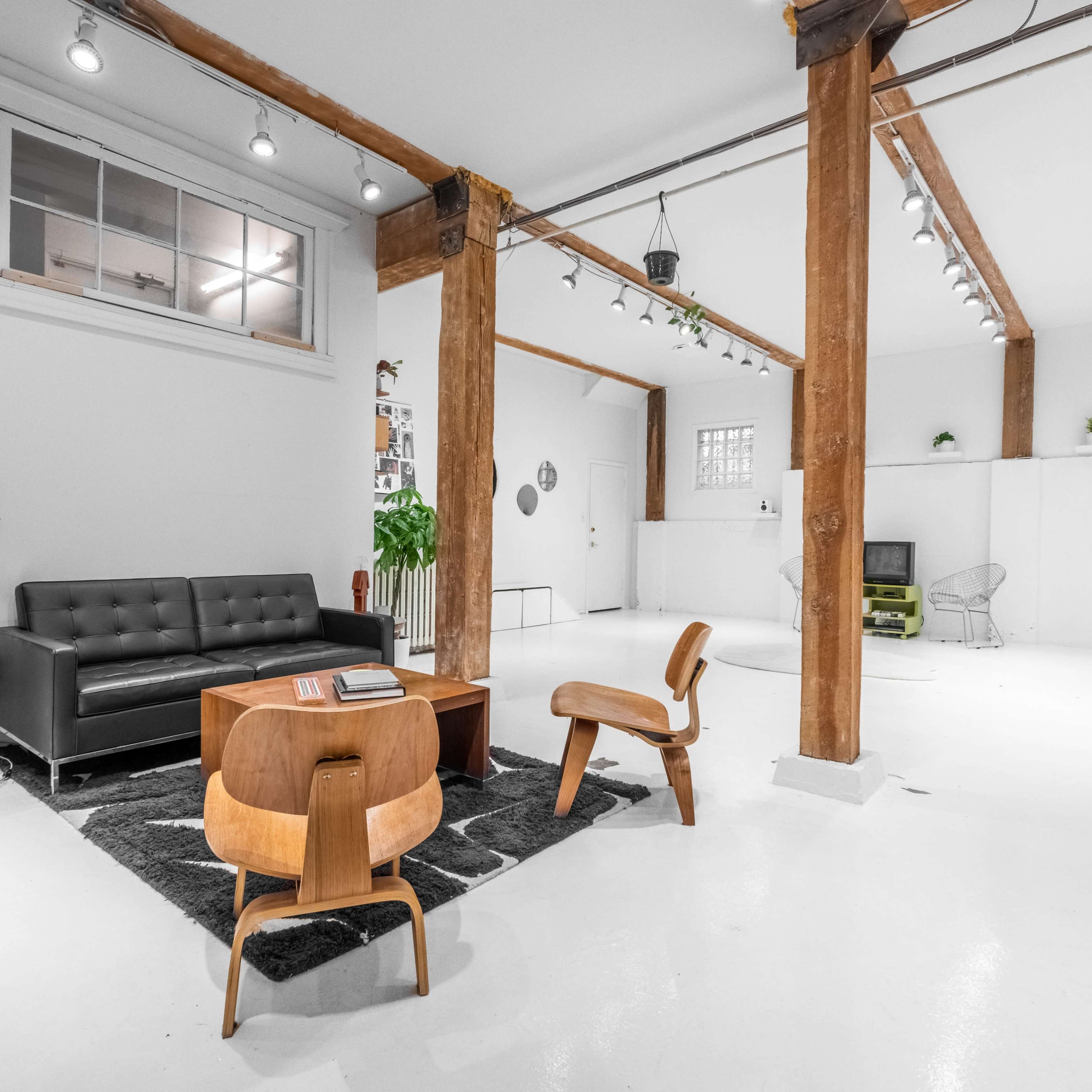 A modern, minimalistic living space featuring a black sofa, a wooden coffee table, and two wooden chairs, illuminated by overhead track lighting and accented by exposed wooden beams.