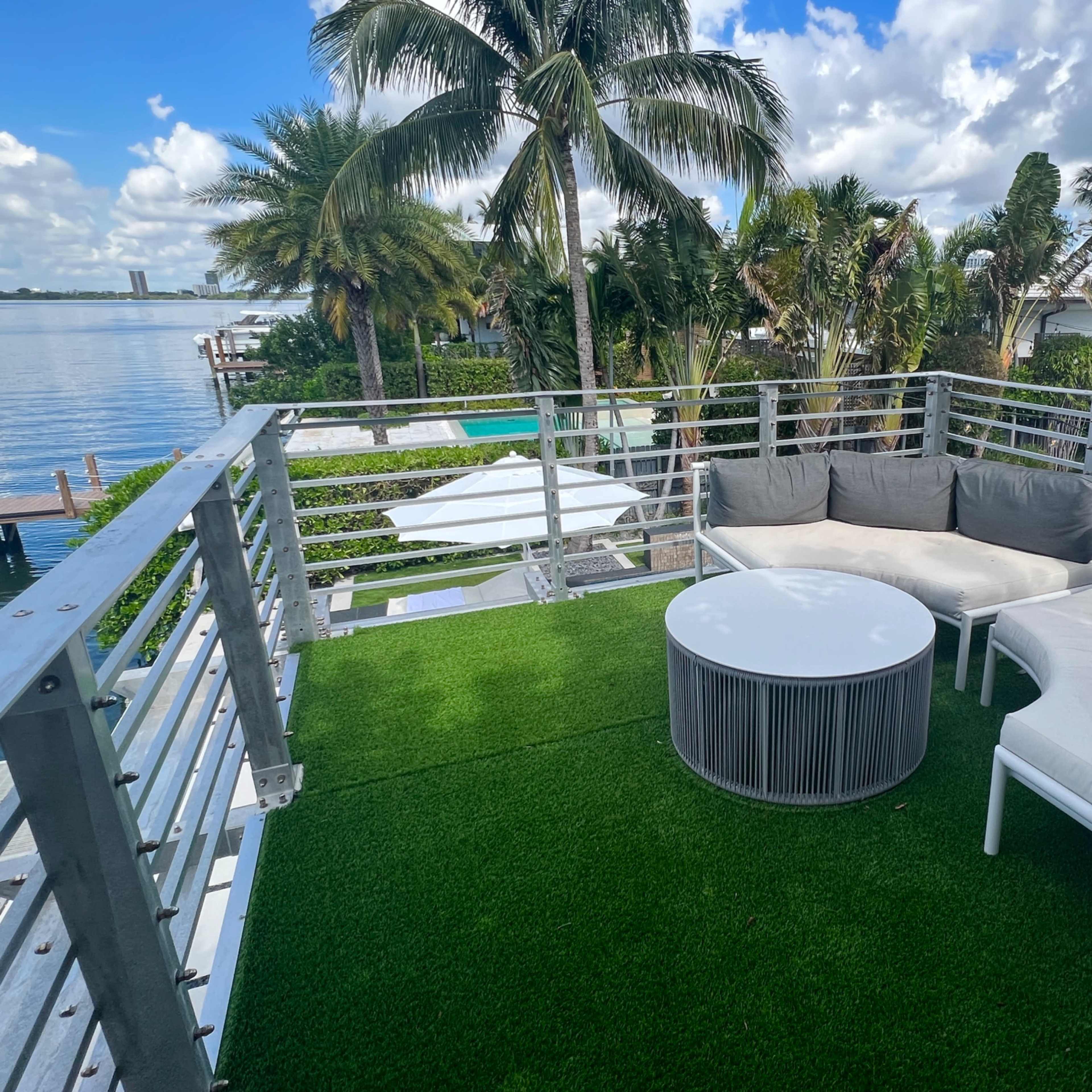 A rooftop terrace features a round table and a white sectional sofa with gray cushions, surrounded by palm trees and a view of the water.