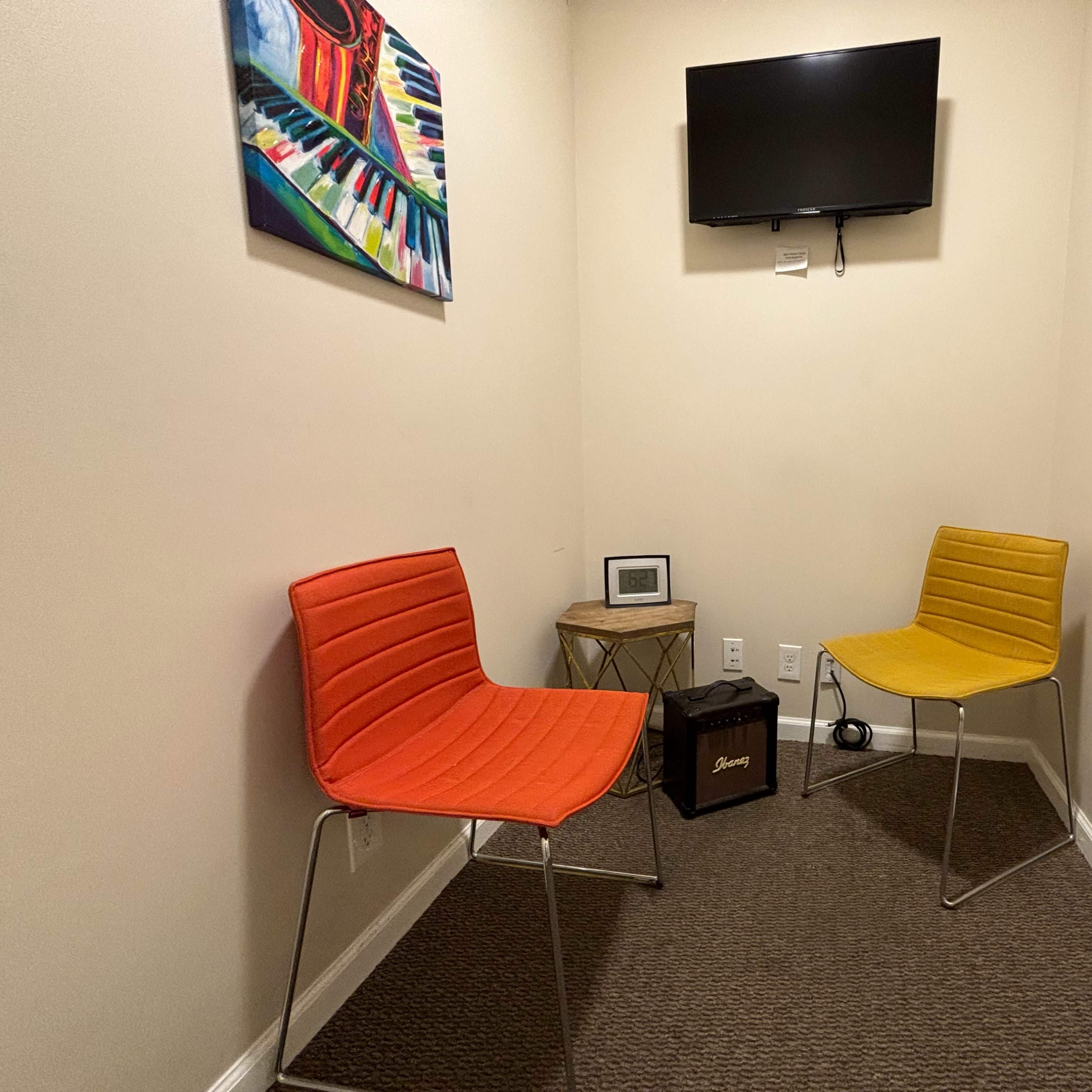 A small room with two chairs, one orange and one yellow, along with a wooden table, a wall-mounted television, and a colorful piano-themed painting.
