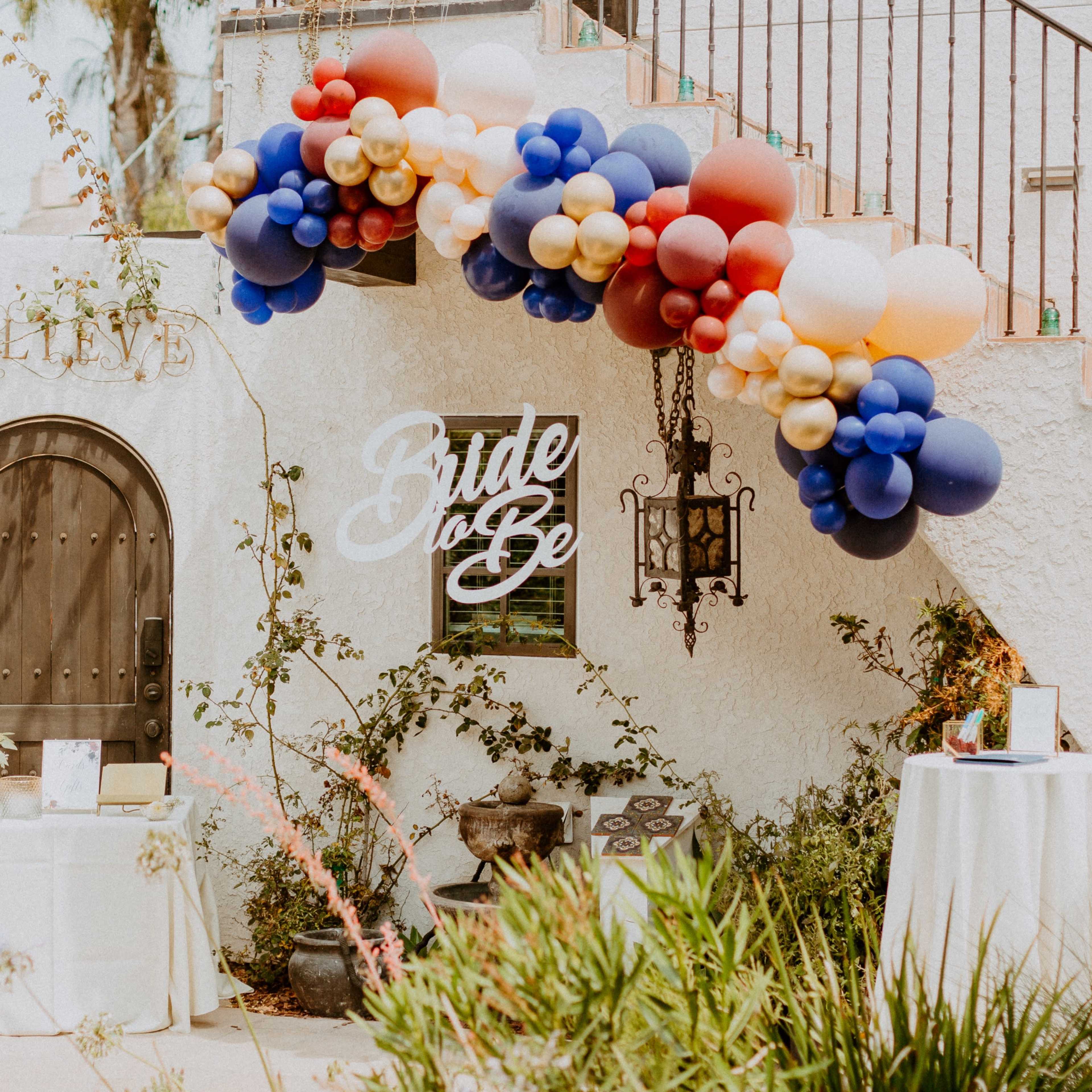 A decorated entrance features a balloon arch in blue, gold, and red, with a sign reading "Bride to Be" alongside tables set for an event.