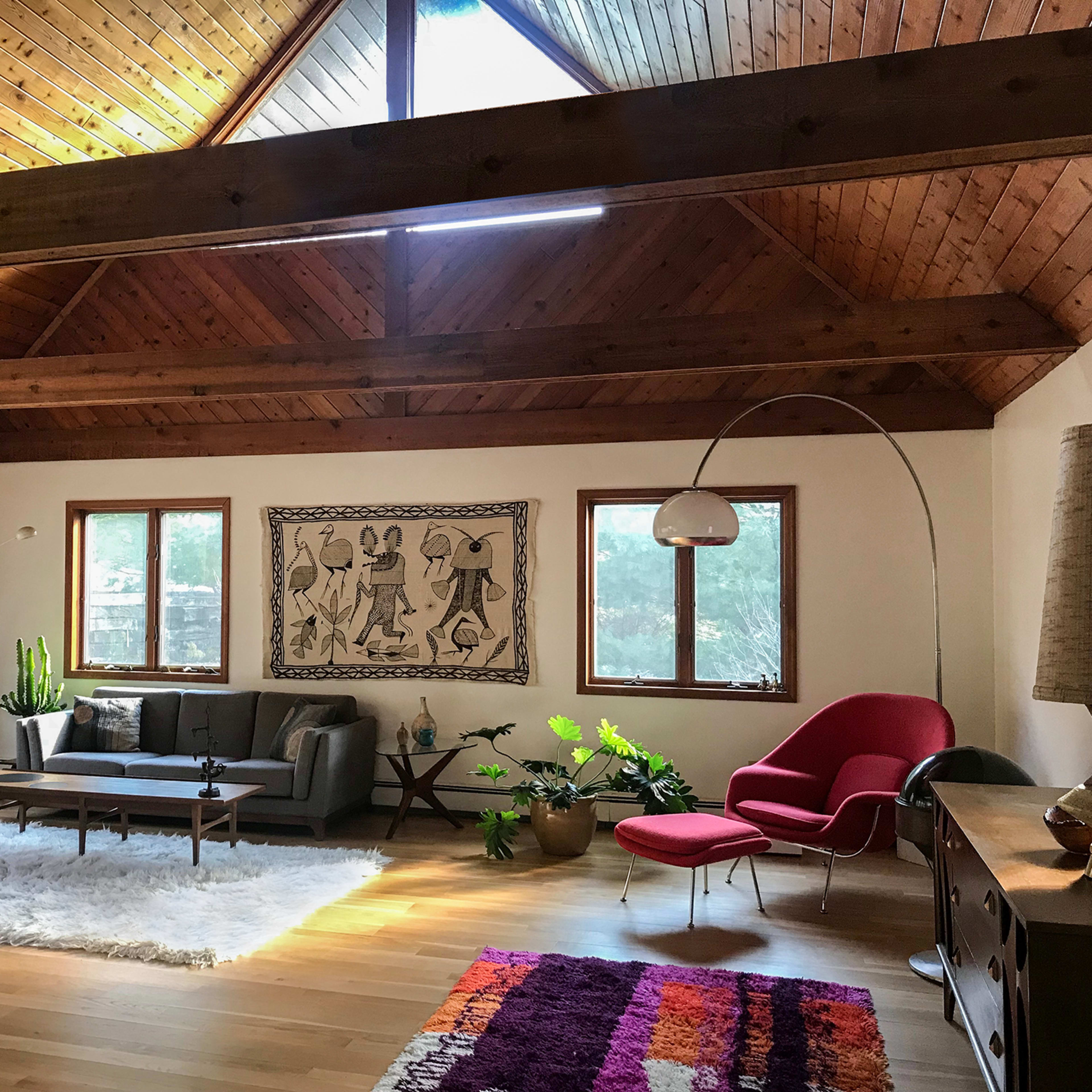 A spacious living room features a high wooden ceiling, large windows, a modern sofa set, and decorative plants.