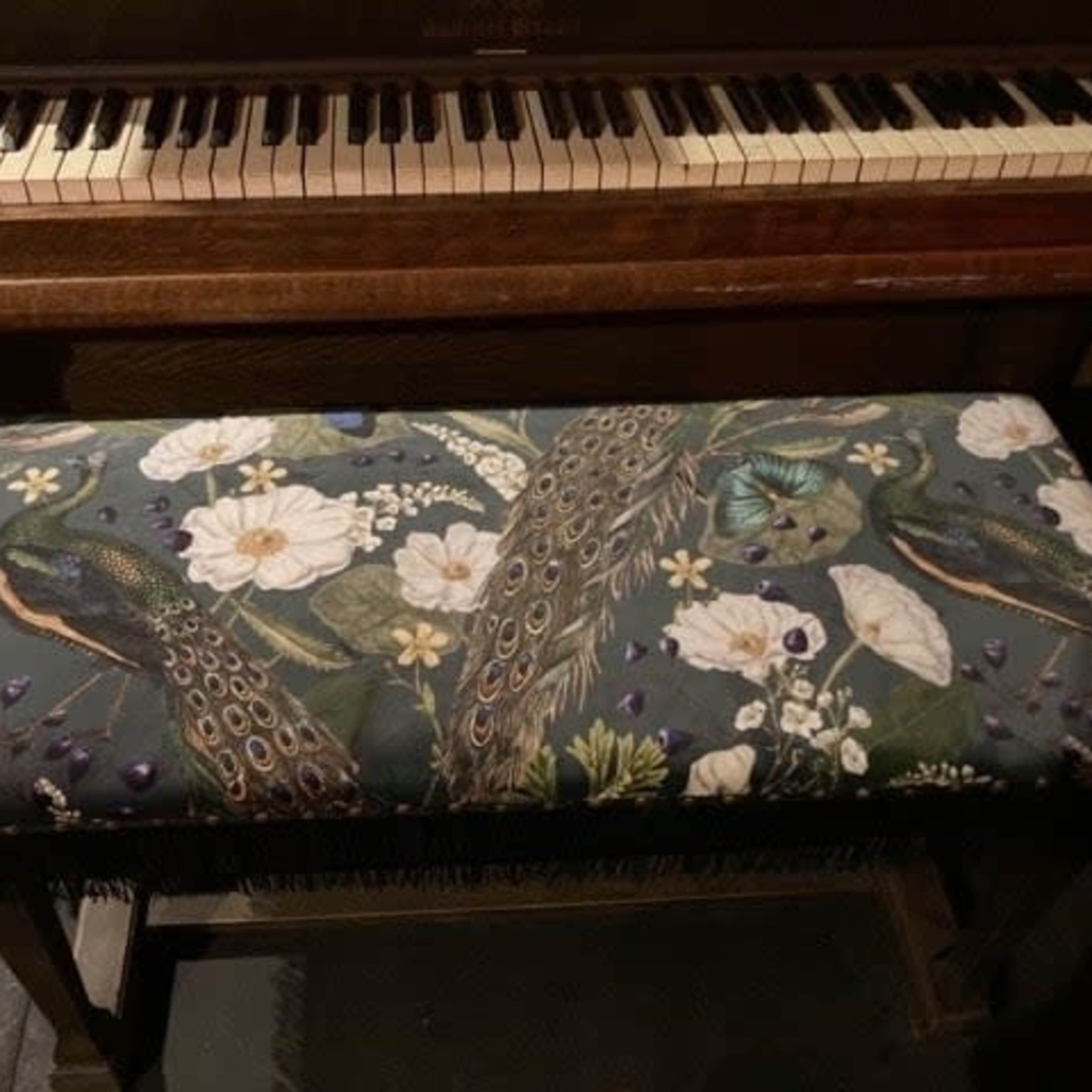 A wooden piano with visible keys is positioned above a decorative bench upholstered with a floral and peacock-patterned fabric.