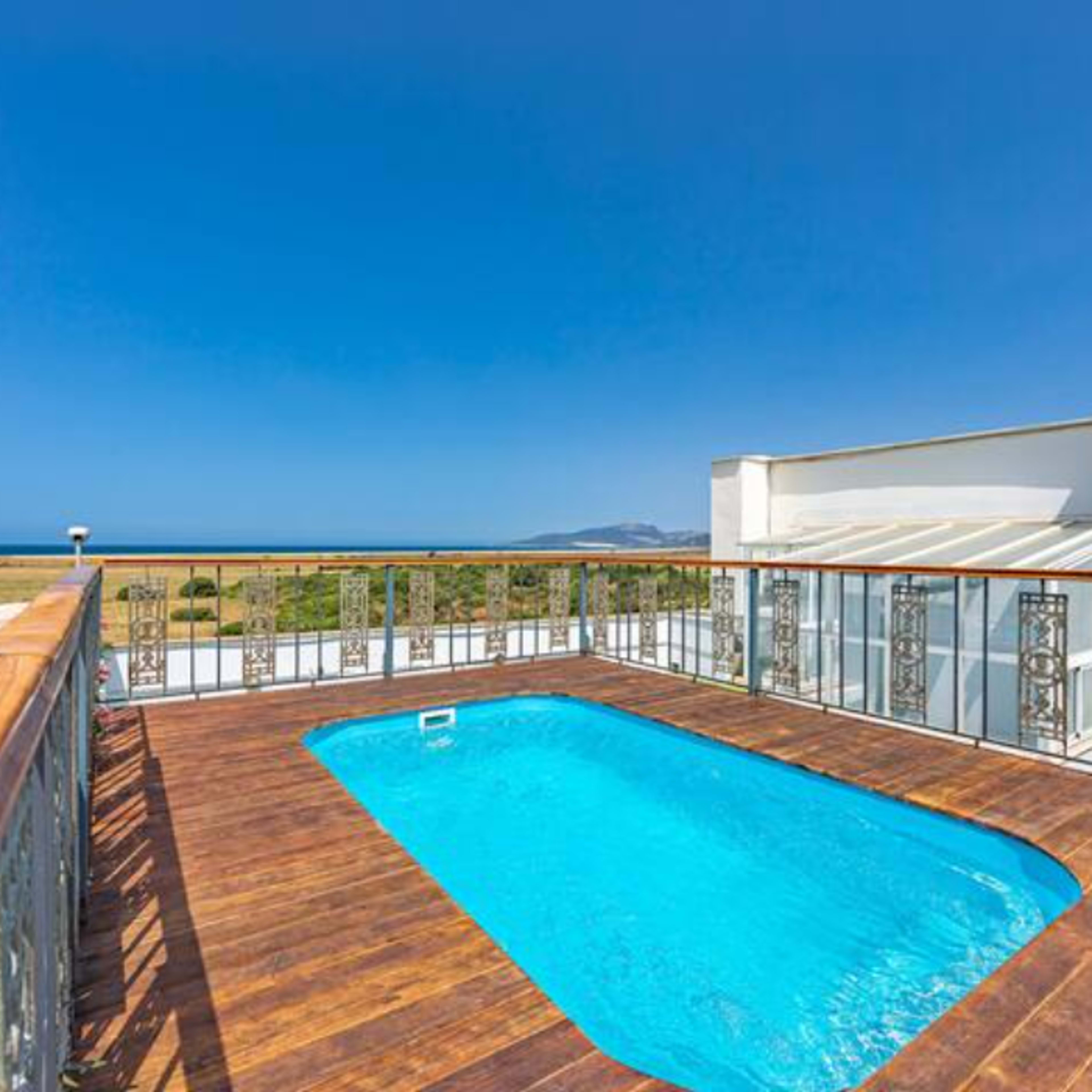 A modern swimming pool surrounded by wooden decking, adjacent to a white building with large glass windows, overlooking a clear blue sea and a grassy landscape.