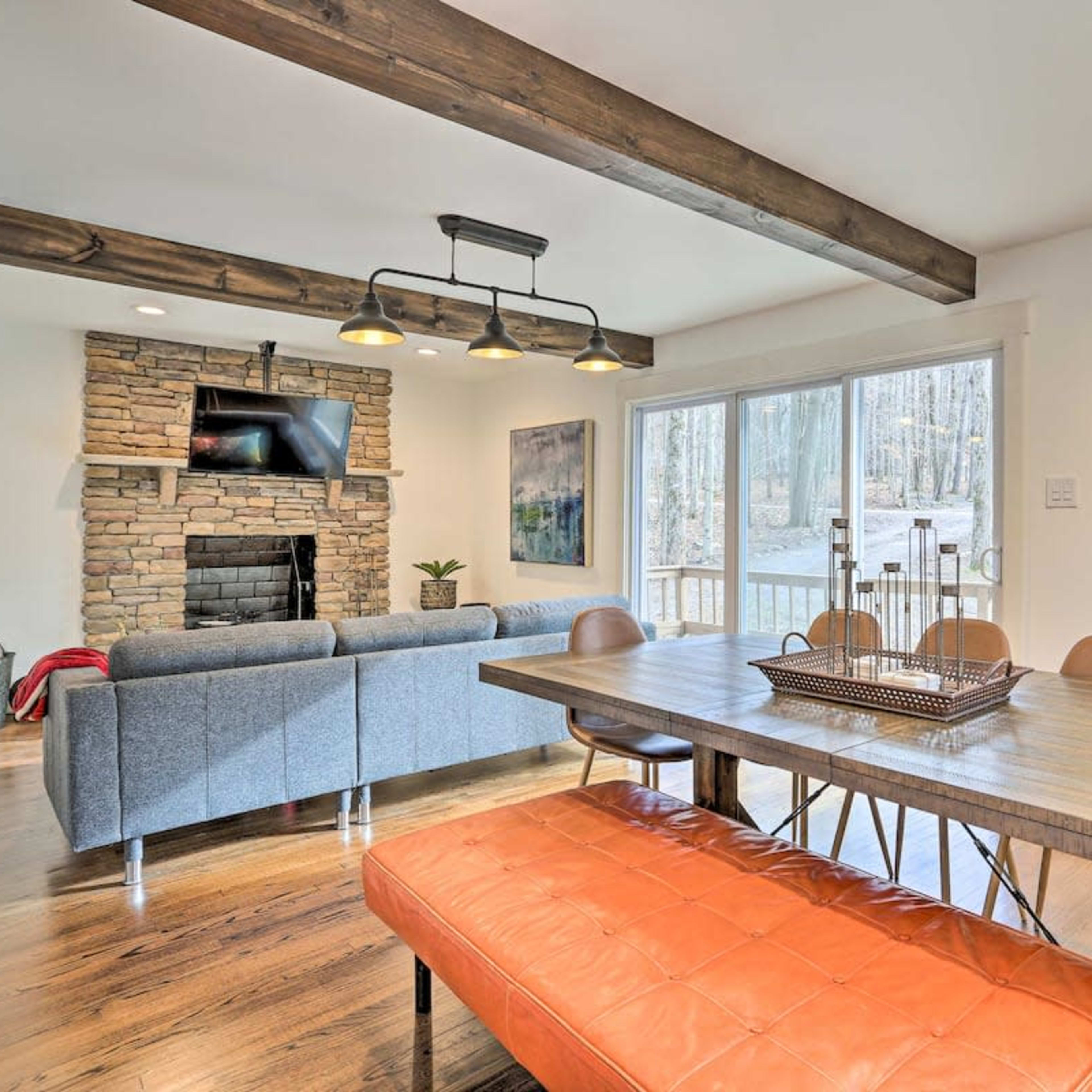 A dining area with a wooden table and orange bench faces a stone fireplace and a large window that offers a view of a wooded area.