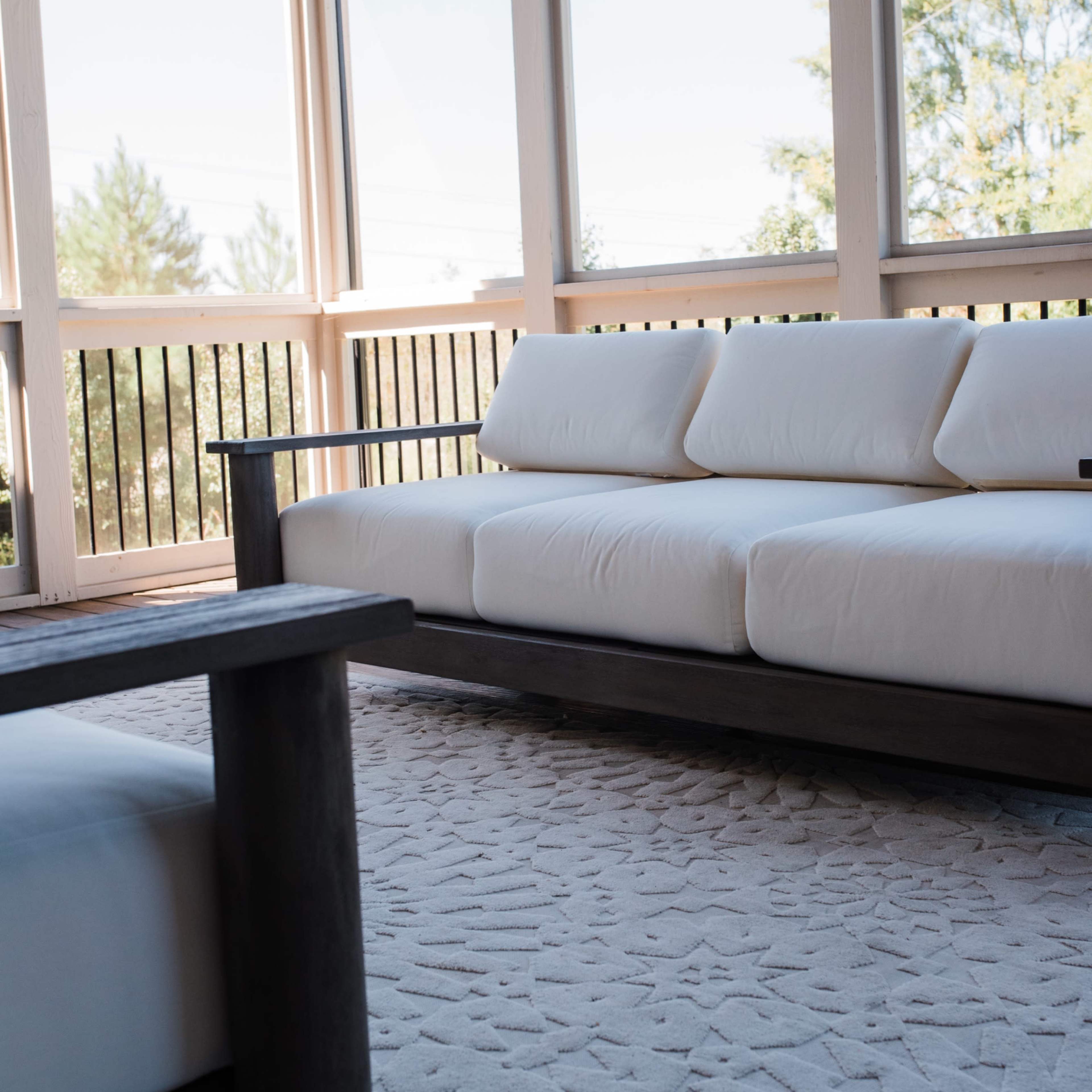 A light-colored sofa is positioned in a sunlit room with large windows, overlooking greenery.