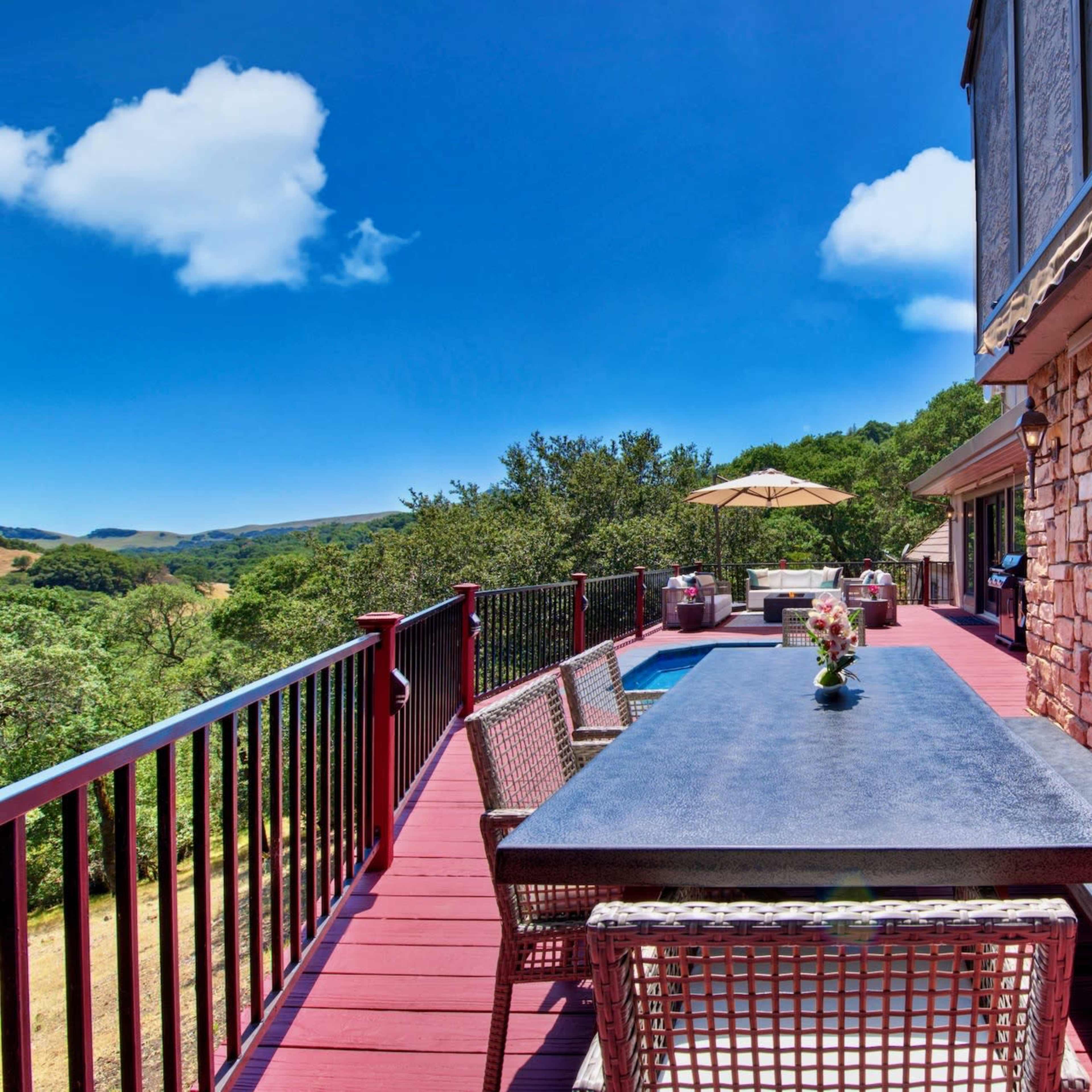 A spacious outdoor deck features a long table surrounded by chairs, overlooking lush green hills and a pool area.