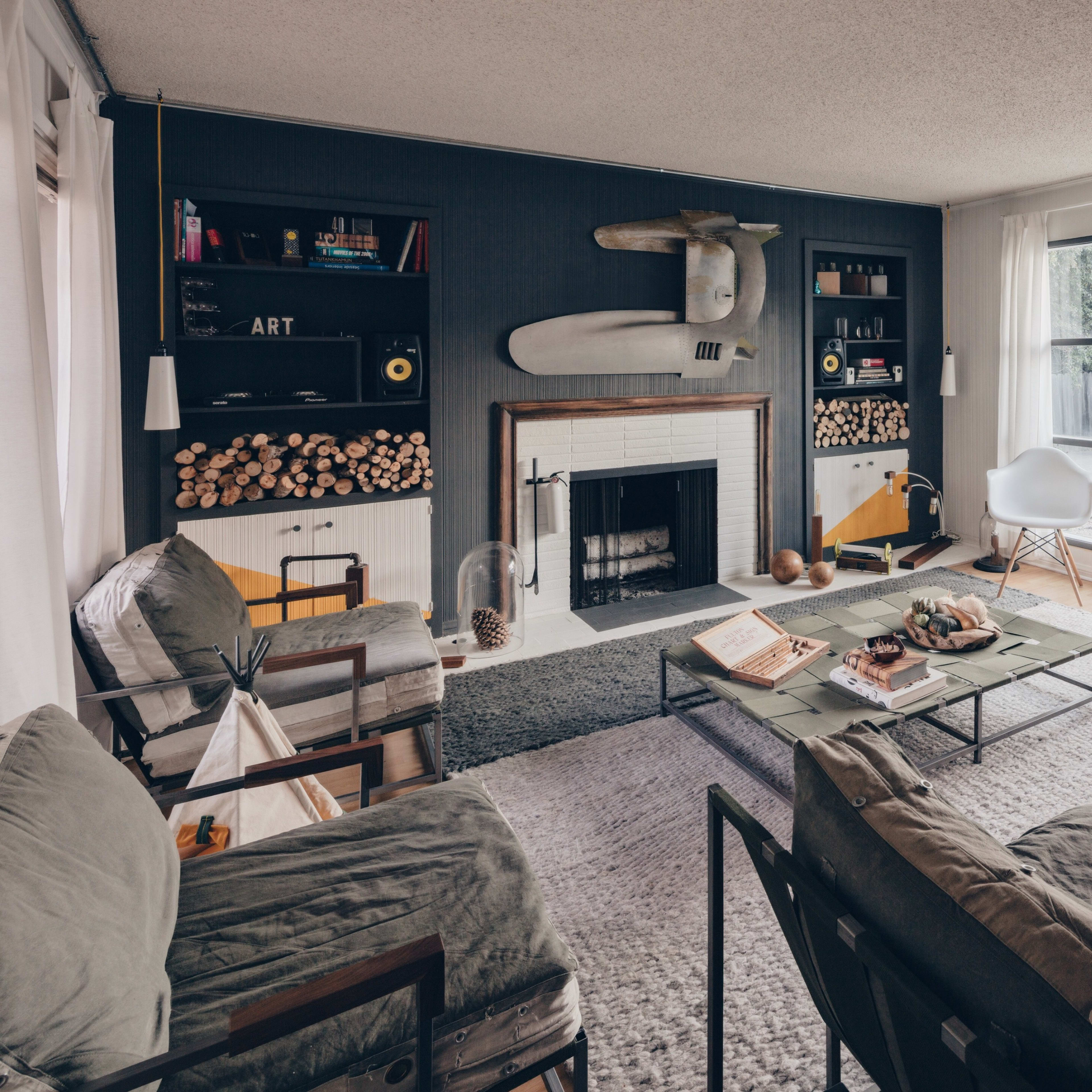 A modern living room features a fireplace flanked by stacked wood, a low coffee table, and large windows providing natural light.