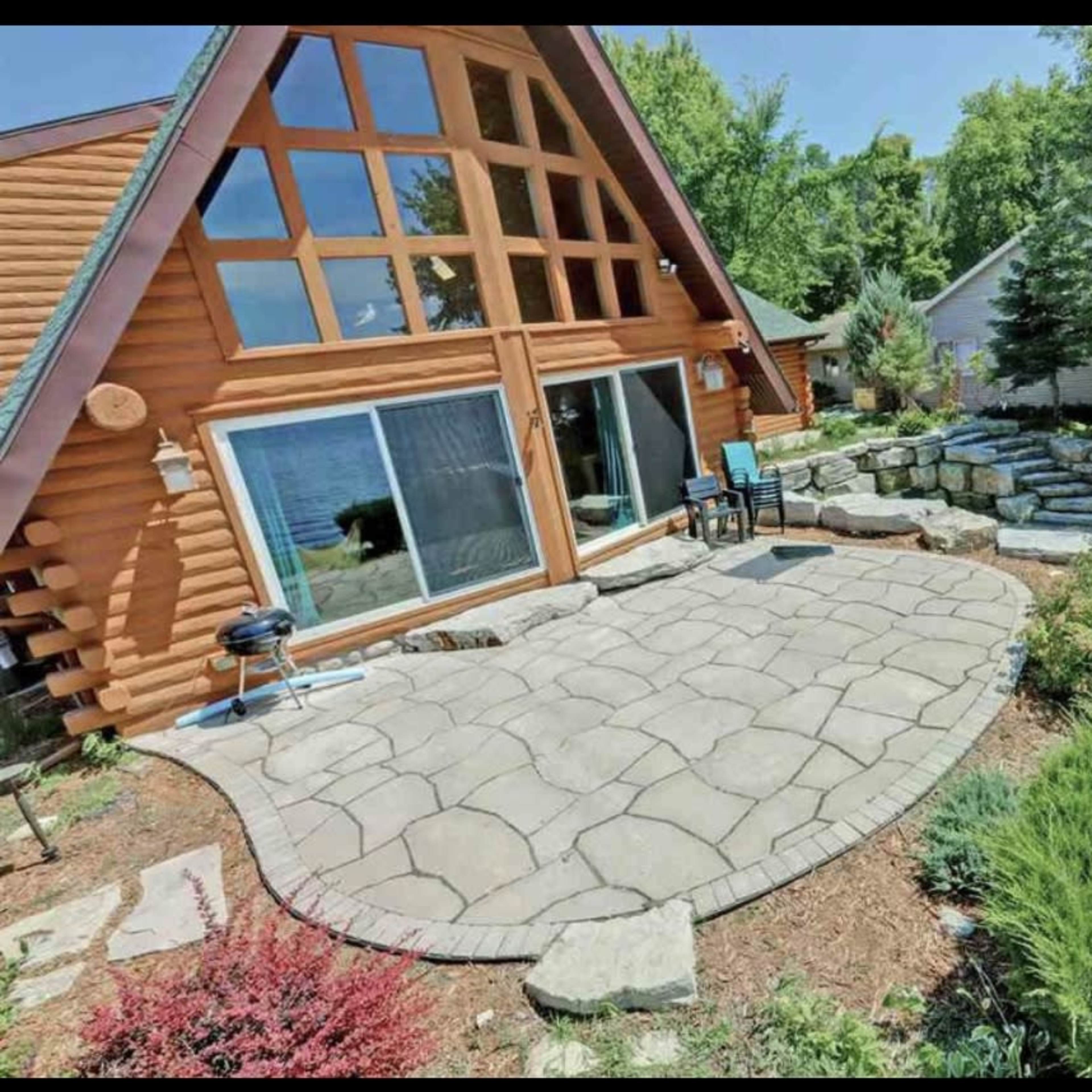 A stone patio with a grill is situated outside a house featuring large windows and an A-frame design, adjacent to a landscaped area with rocks and greenery.