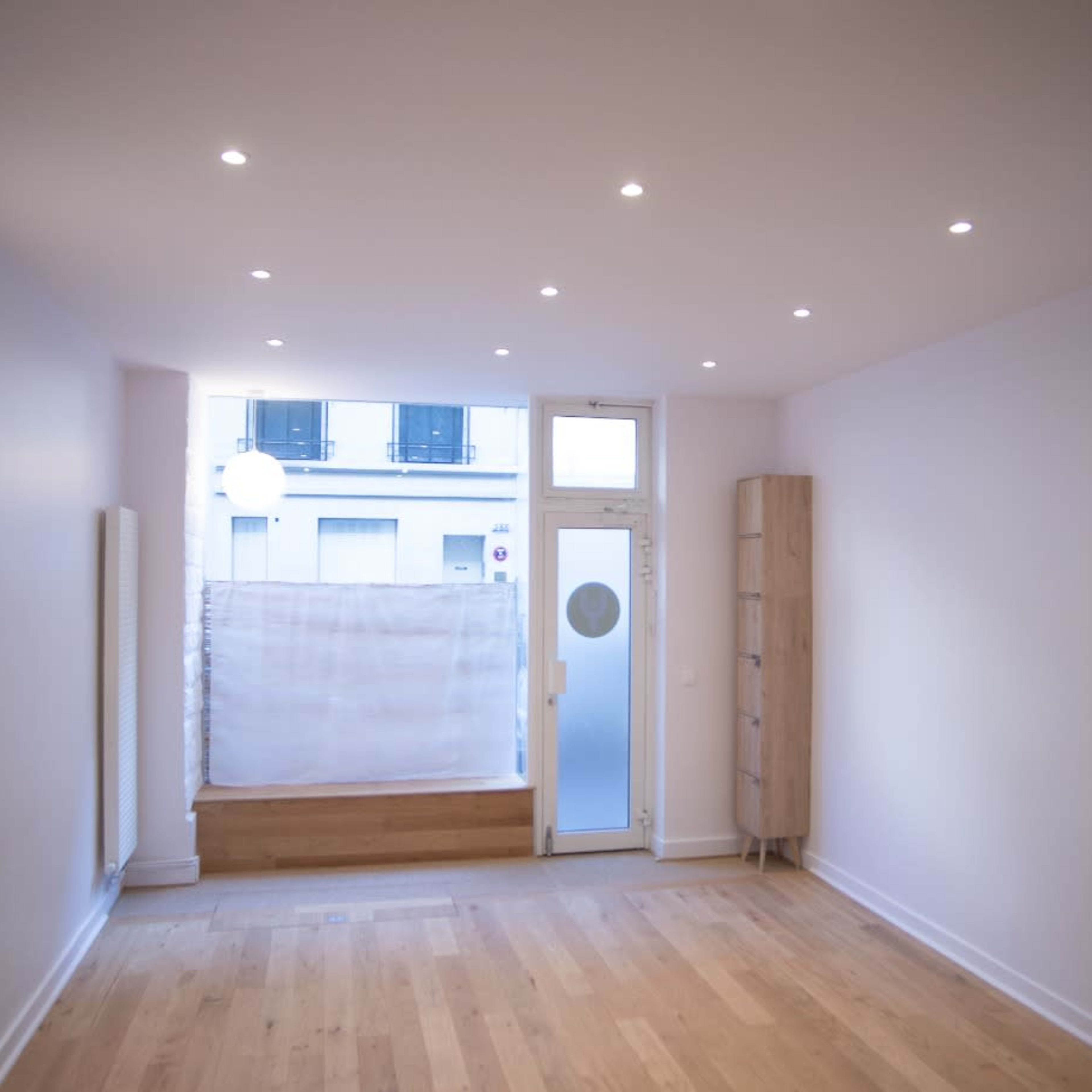 An empty room with wooden flooring, a large window with a partially drawn curtain, and a simple wooden cabinet against one wall.