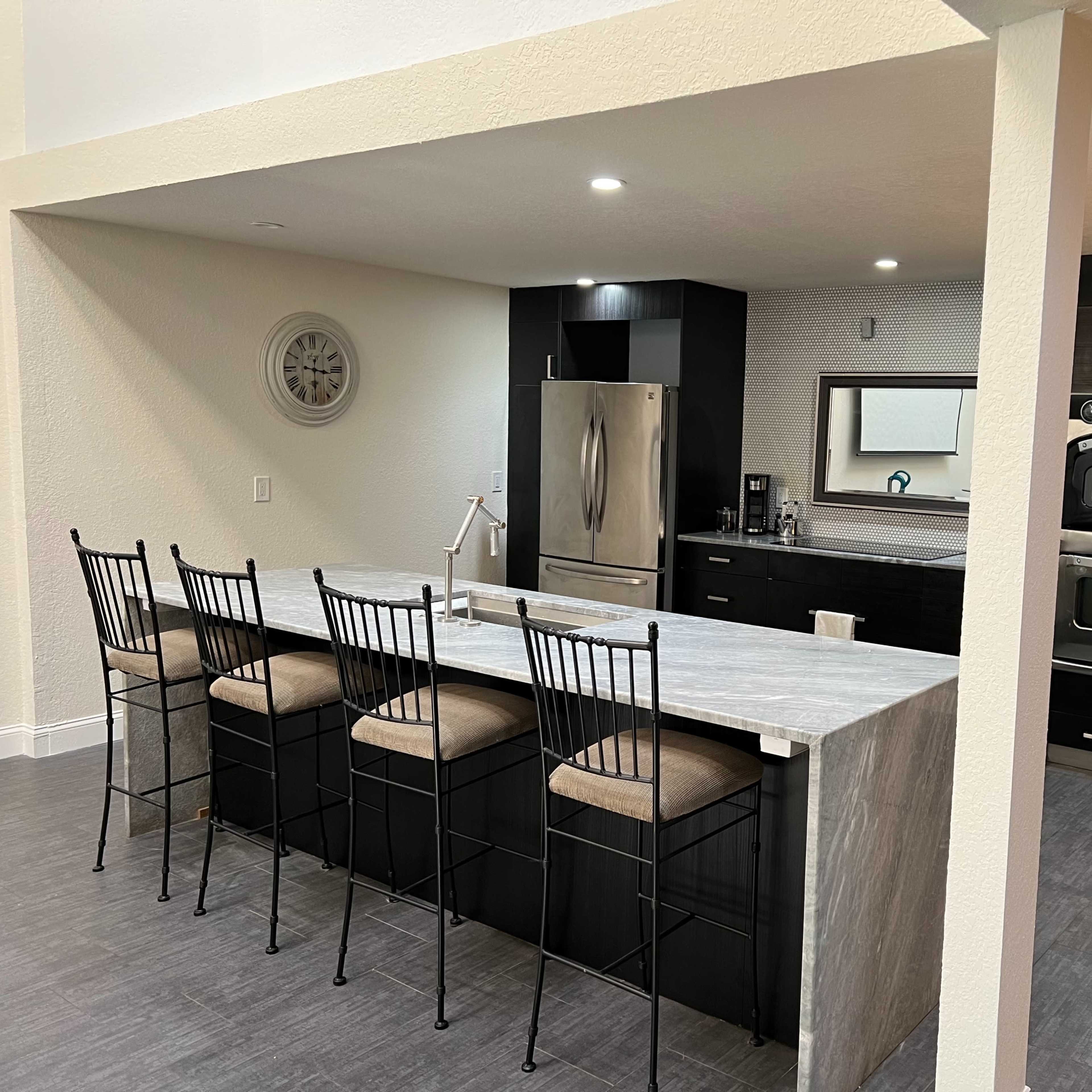 A modern kitchen with a long, gray stone island and black cabinetry, featuring a stainless steel refrigerator and an oven.